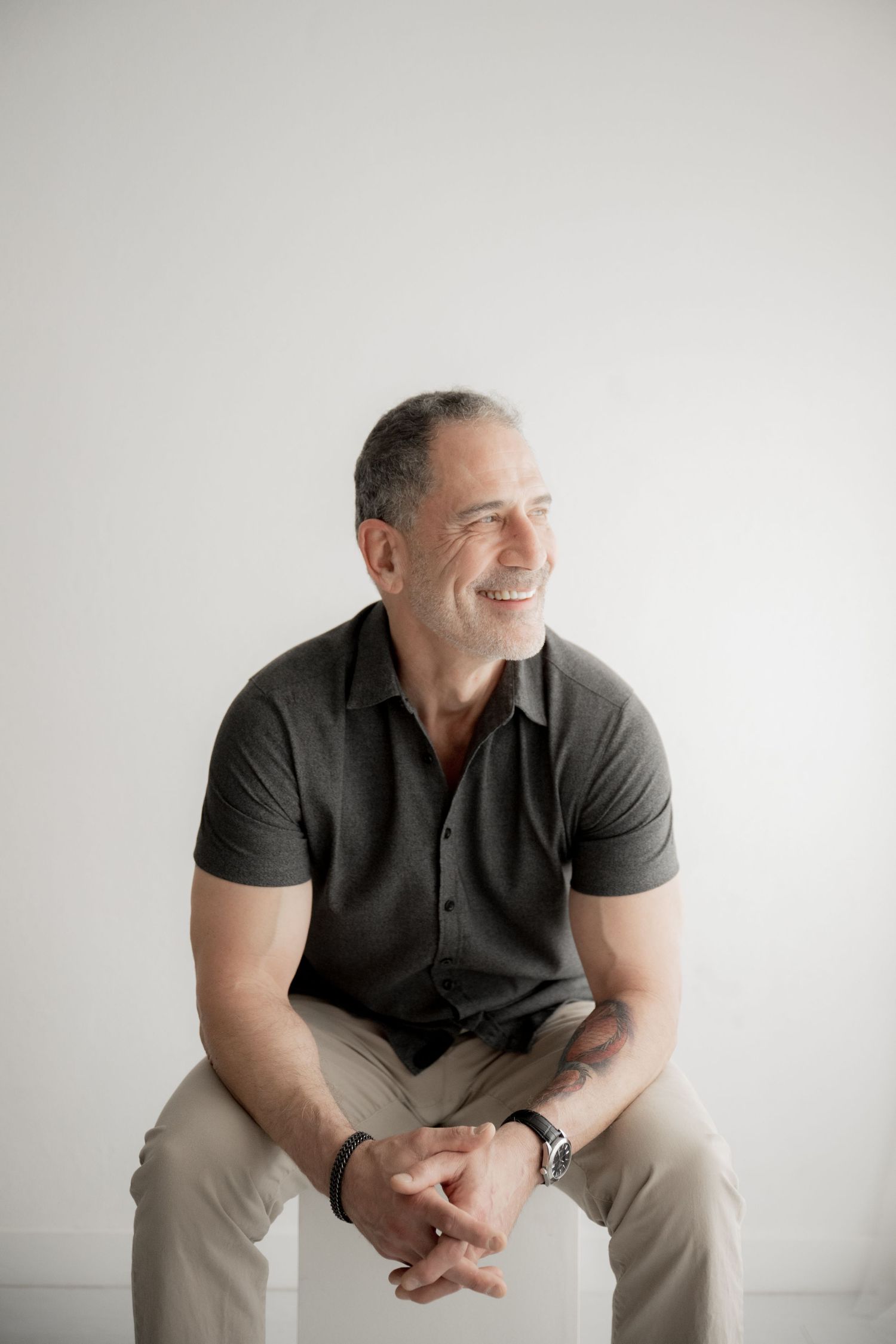 A casually dressed person sits on a white chair against a light background, smiling warmly to the side.