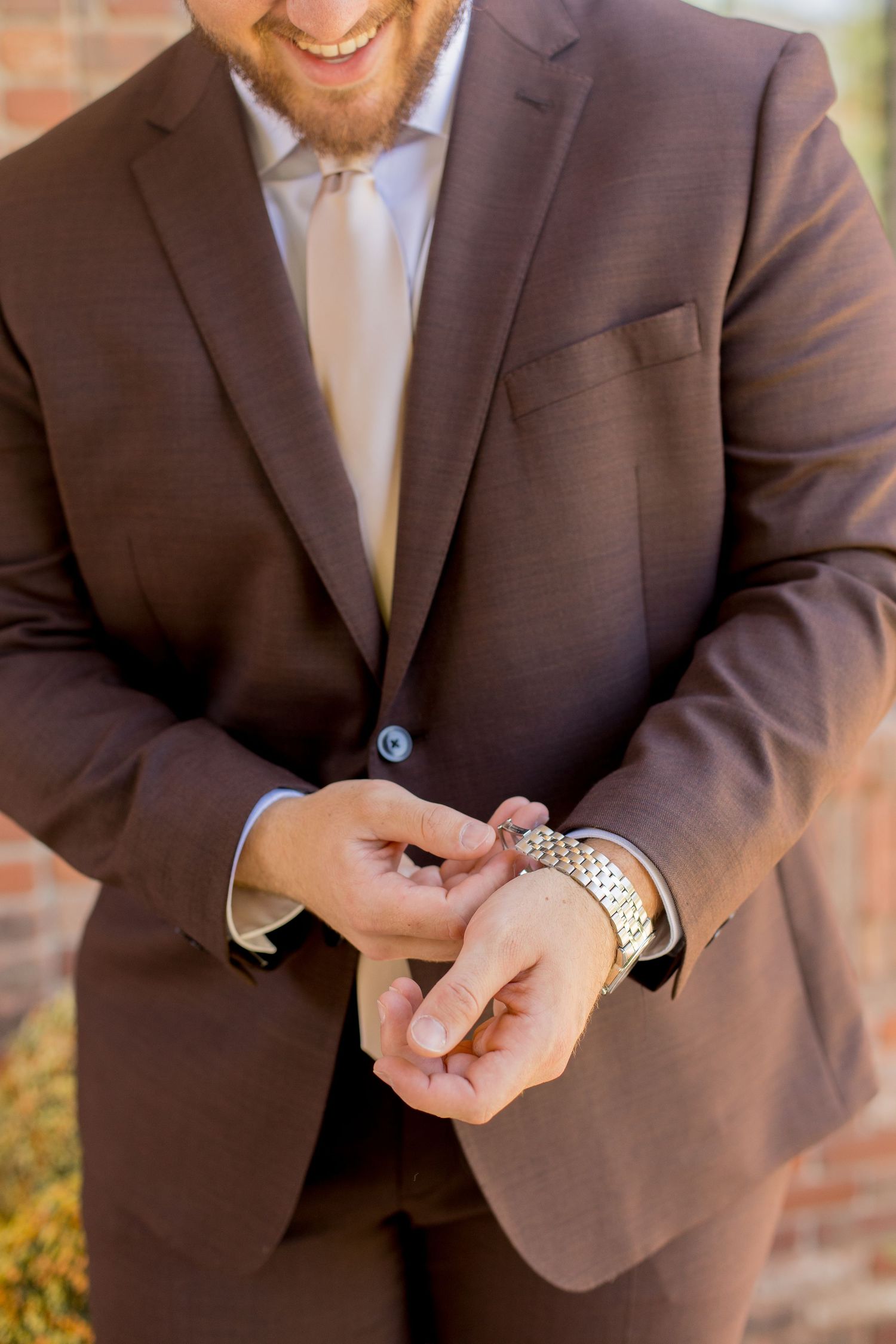 Close-up view of someone in a brown suit jacket checking their silver wristwatch in warm sunlight.