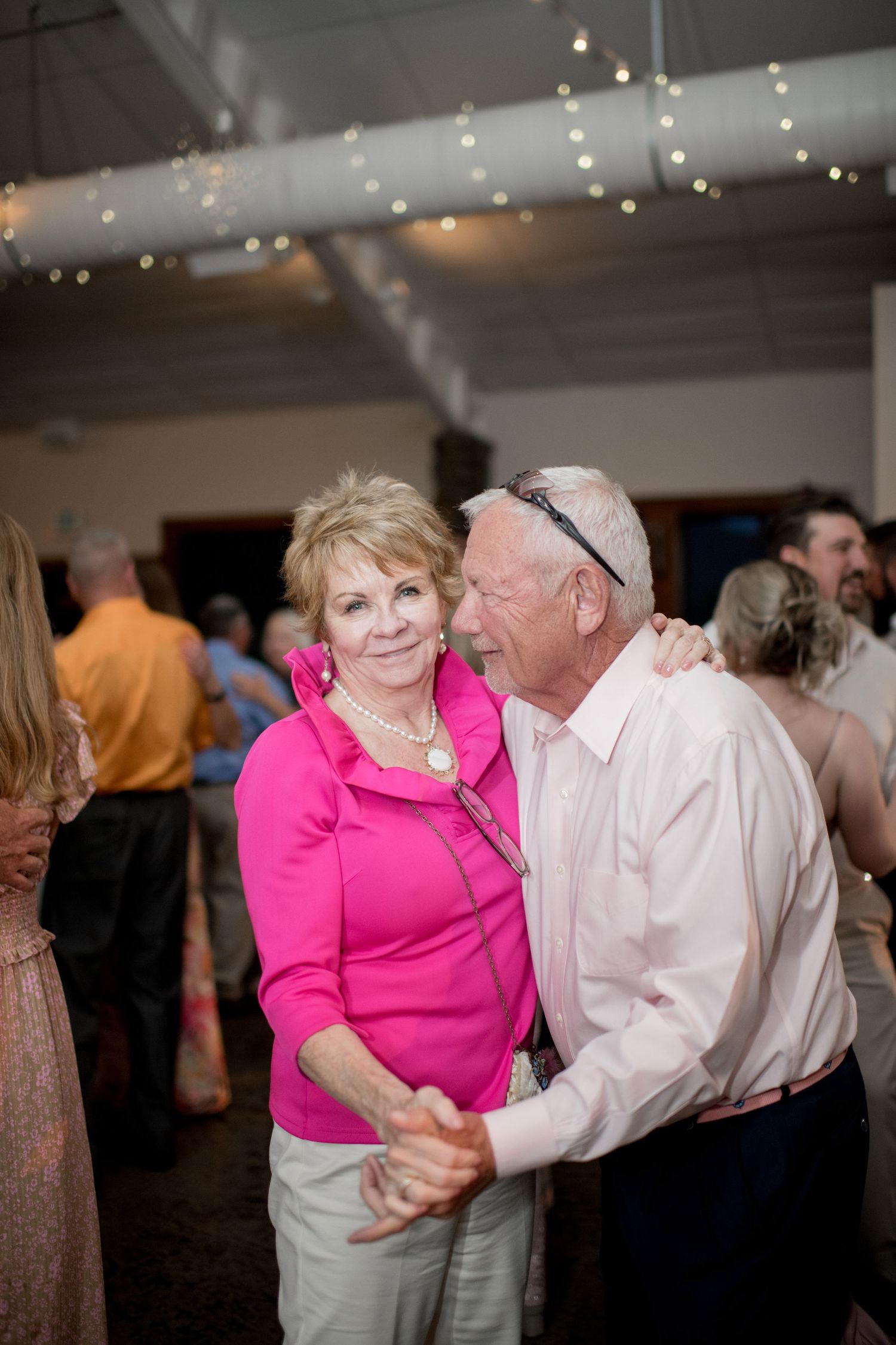 Couple dancing together at a wedding reception with string lights overhead, woman in bright pink top and man in white shirt.
