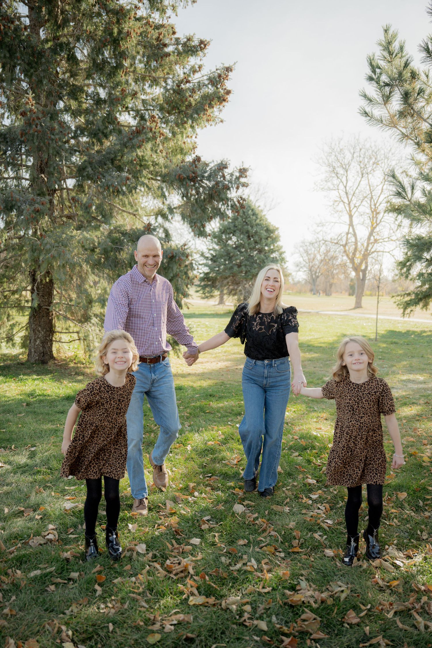 A family plays together in autumn leaves under tall evergreen trees on a sunny afternoon.