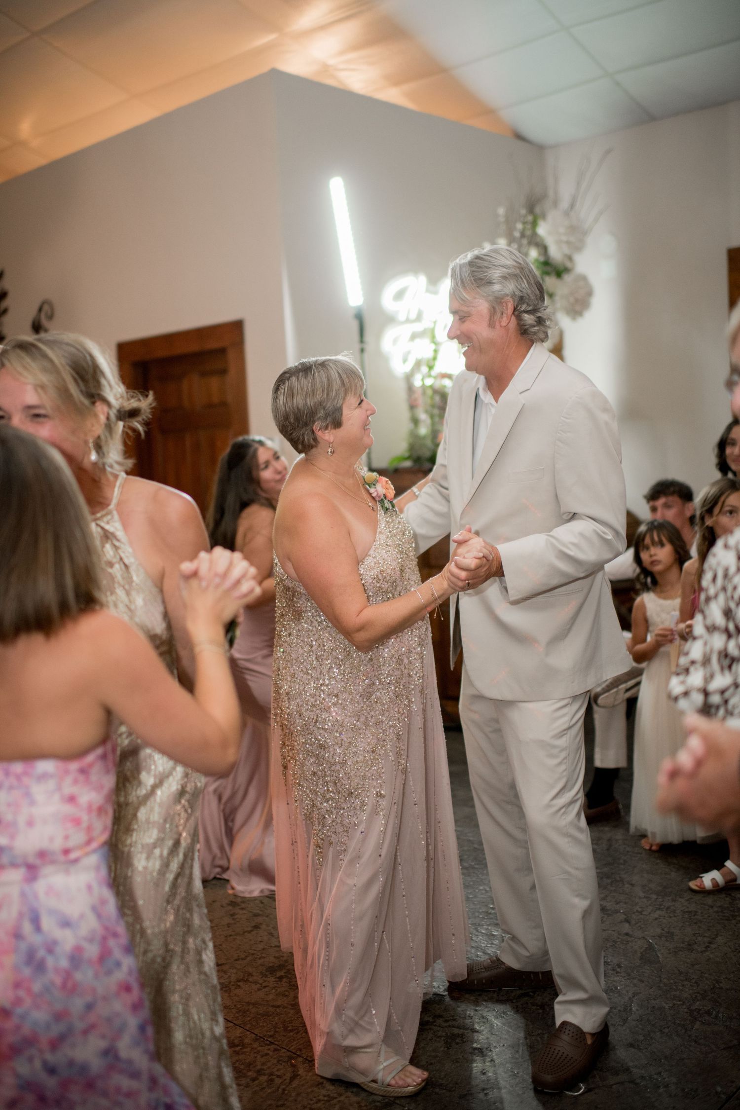 Wedding guests dancing together at a reception in a bright indoor venue with floral decorations.