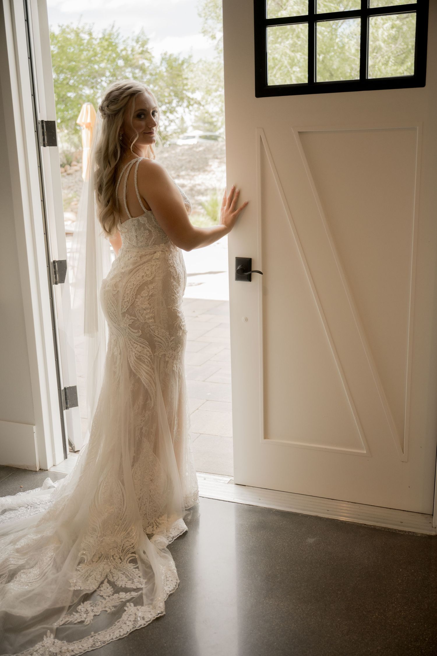 A bride in a flowing lace wedding gown stands elegantly by a white doorway in natural light.