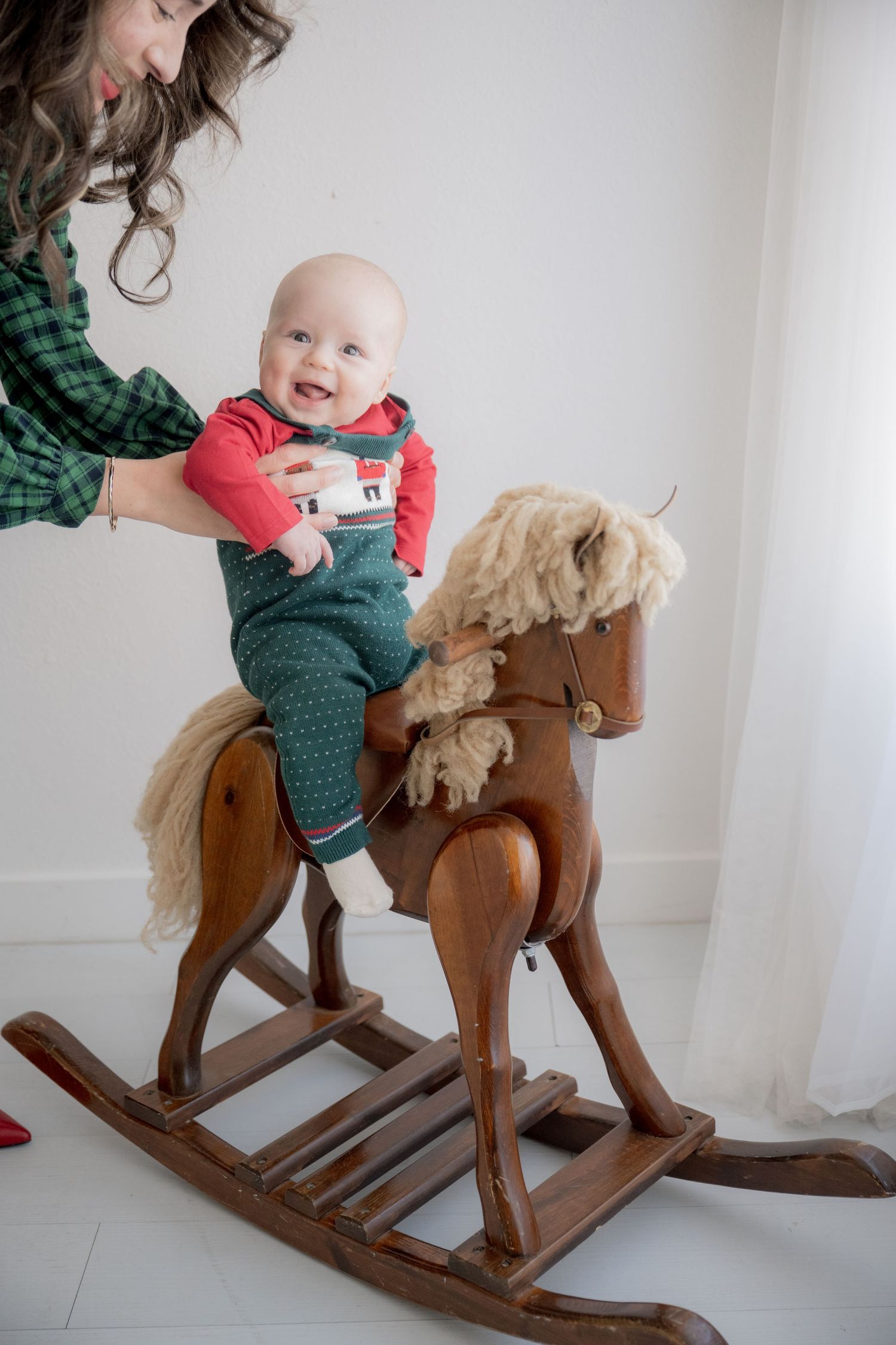 Child in green shirt enjoys playtime on a wooden rocking horse toy against white wall.