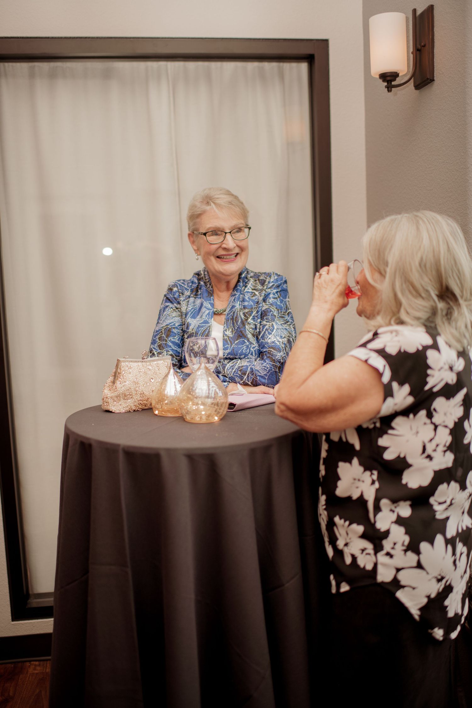 Two women chat at a small round table with dark tablecloth and glassware in an indoor setting.
