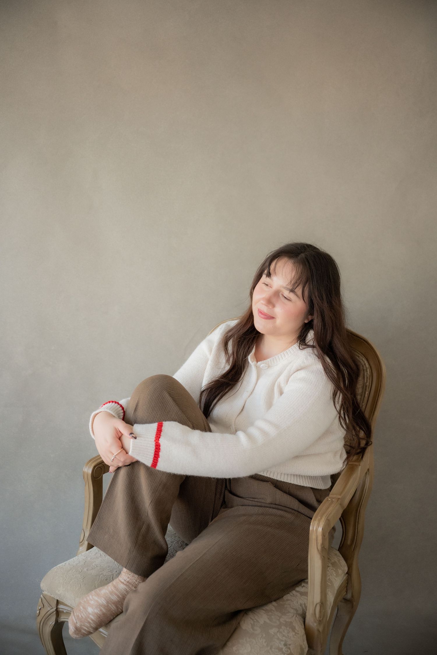 A person in cozy white sweater and brown pants relaxes in an antique chair against a neutral wall.