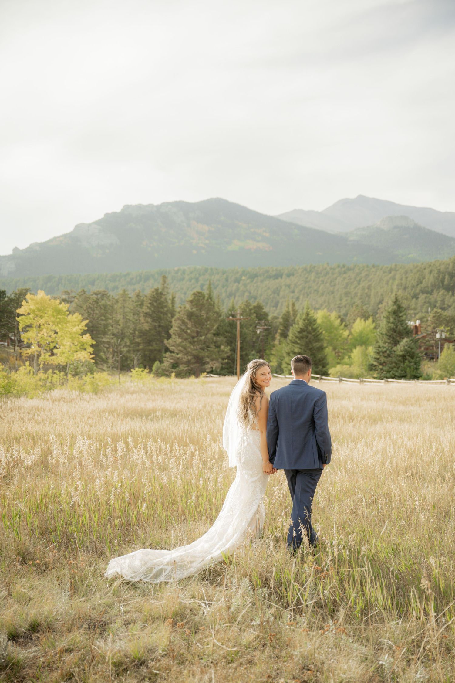 A couple walks hand in hand through a golden mountain meadow with evergreen trees and misty peaks in the background.