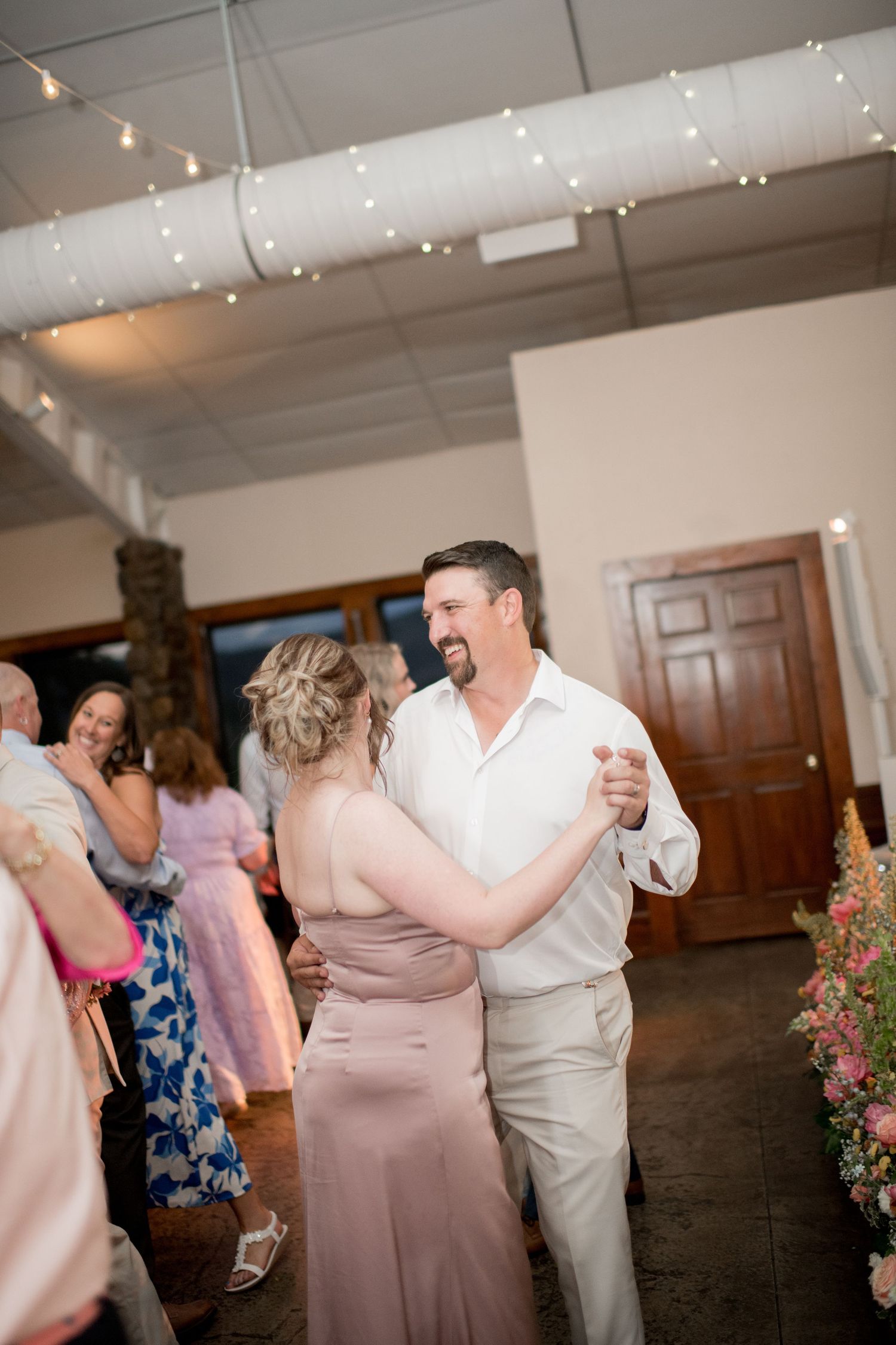 Wedding guests dancing and celebrating together at a reception with floral decorations and warm lighting.