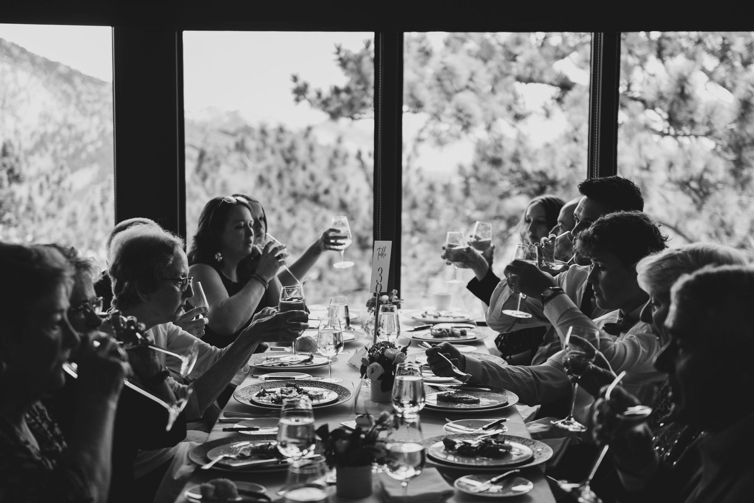 Black and white photo of a dinner party gathering with people seated at a long table by large windows overlooking nature.