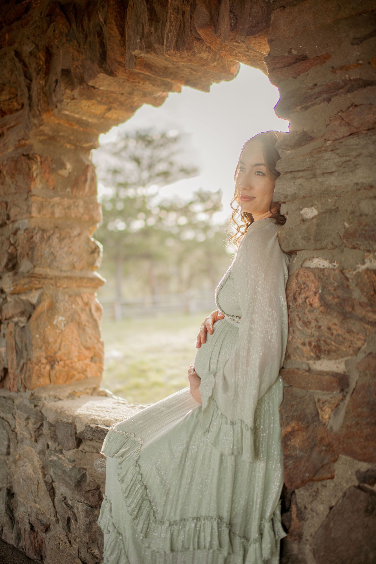 A dreamy portrait taken through a stone archway shows a figure in a flowing sage green vintage-style dress at sunset.