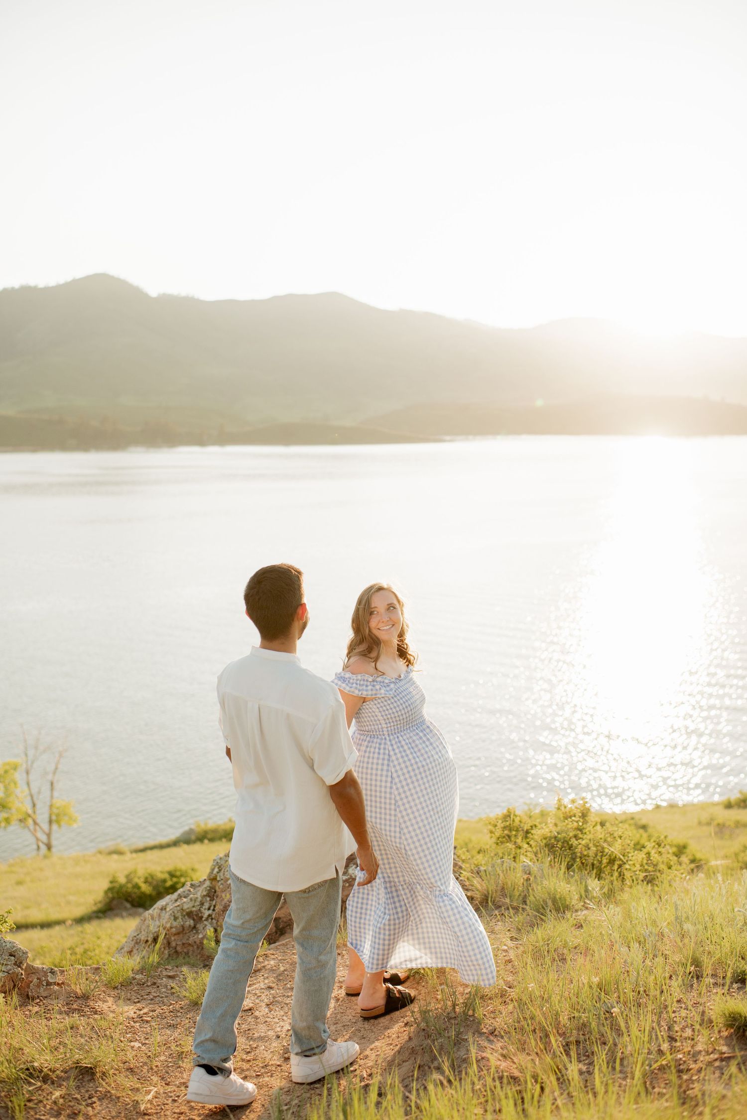 Couple sits together on grassy hillside overlooking scenic lake at sunset.
