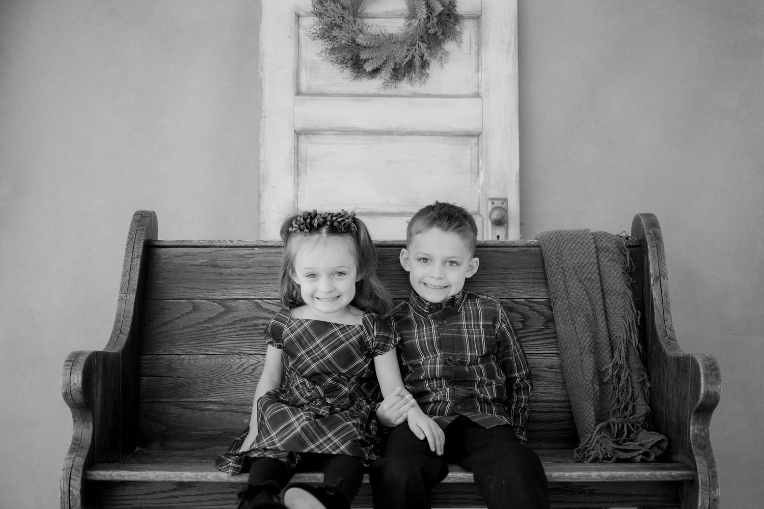 A black and white photo of two young children sitting together on a wooden church pew beneath a decorative wreath.