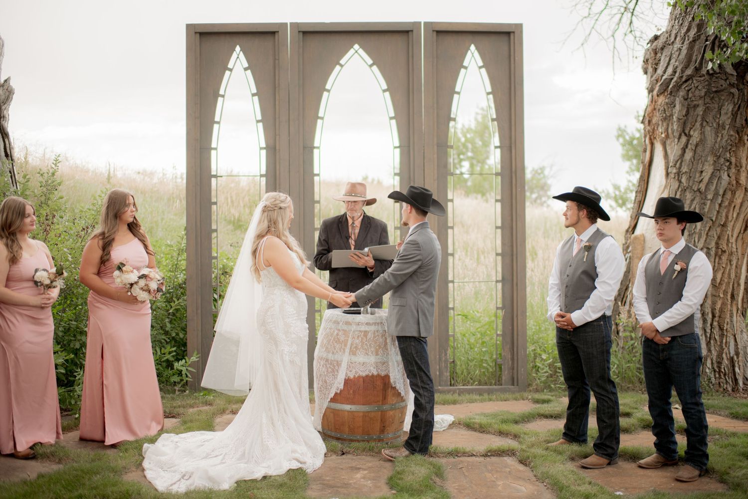 Wedding ceremony takes place under elegant stone arches with bridal party in pink dresses and groomsmen in gray vests.