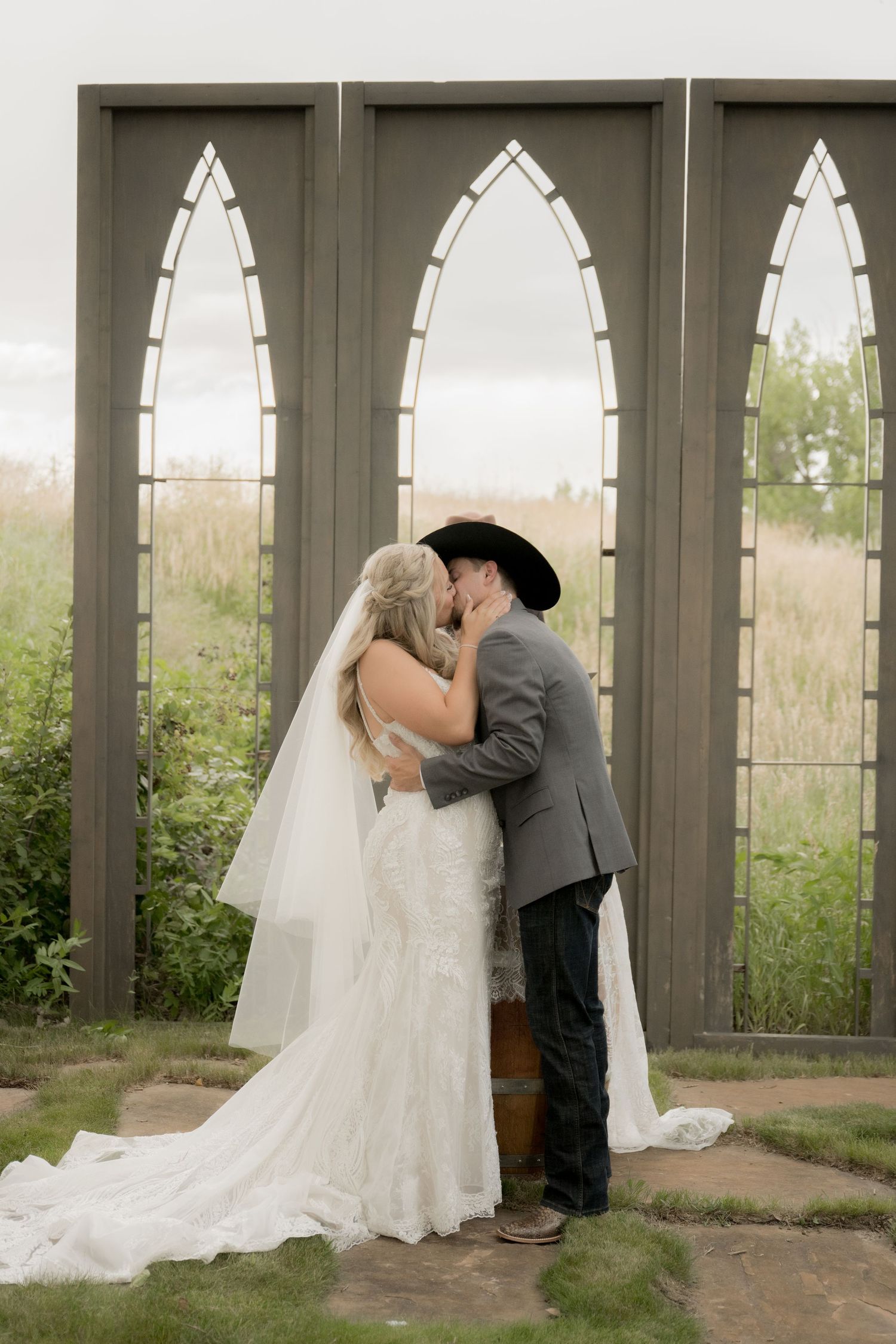 A romantic wedding couple shares a tender kiss in a rustic chapel setting with arched windows.