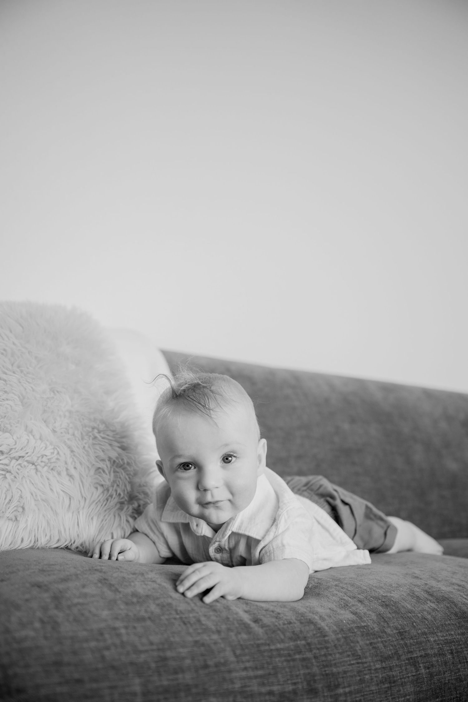 A young child in light colored clothing lies on a couch with a fuzzy cushion in black and white photography.