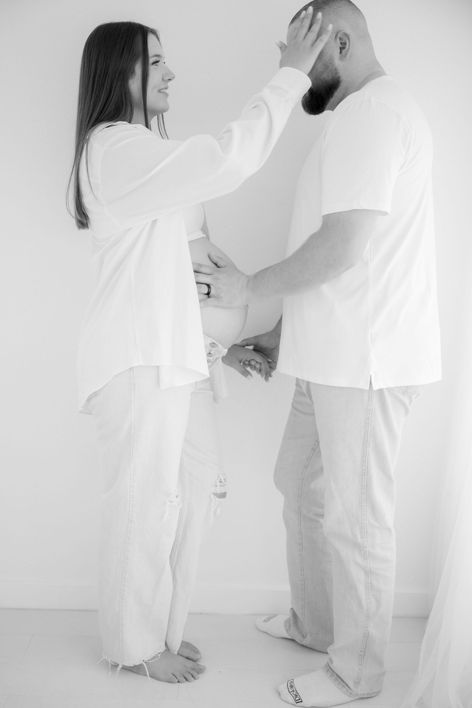 Two people dressed in all white stand facing each other against a white background in an intimate artistic pose.