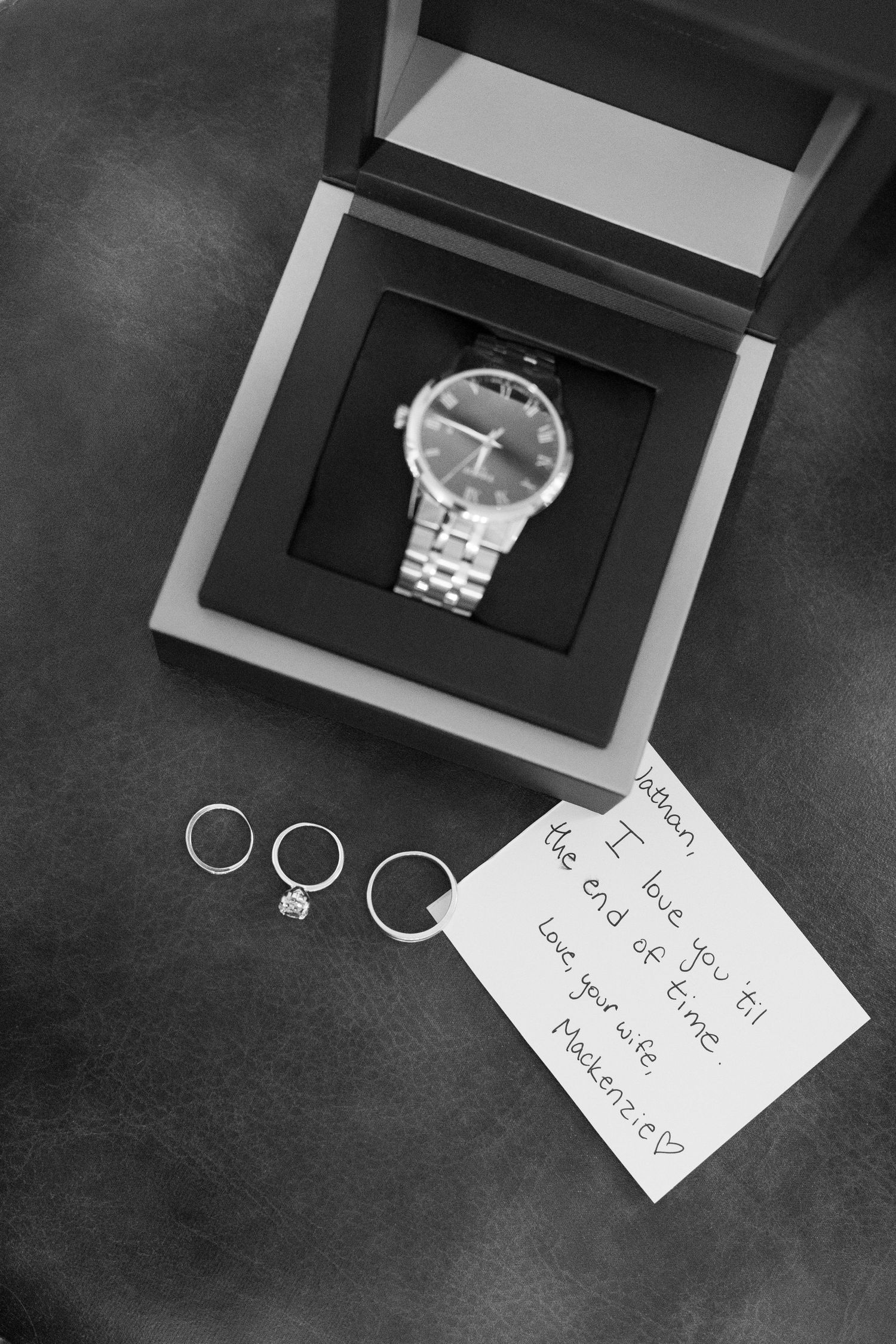 Black and white photo of a watch in a gift box with rings and a handwritten note below.