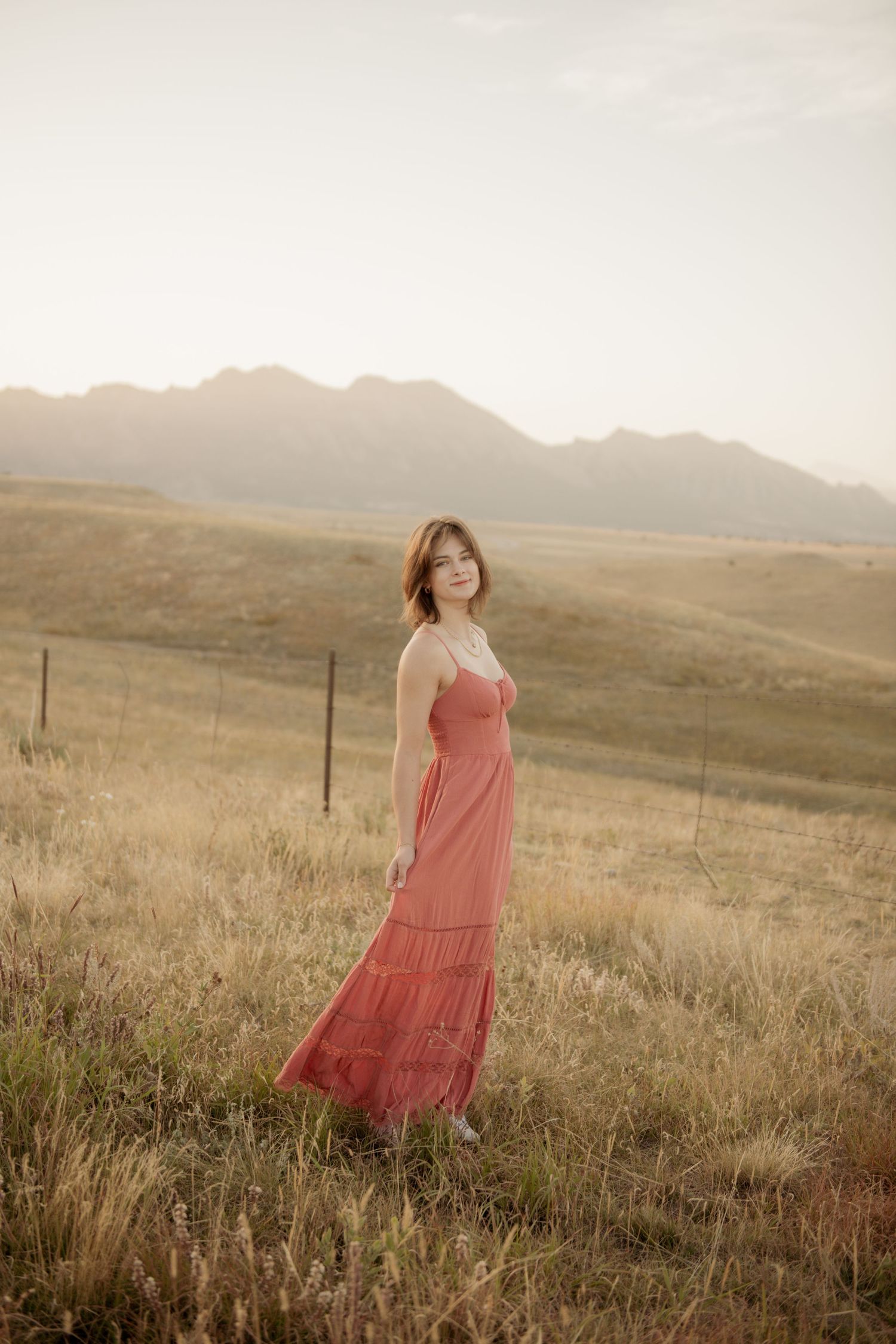 A figure in a flowing pink dress stands in a golden prairie field at sunset with mountains in the distance.