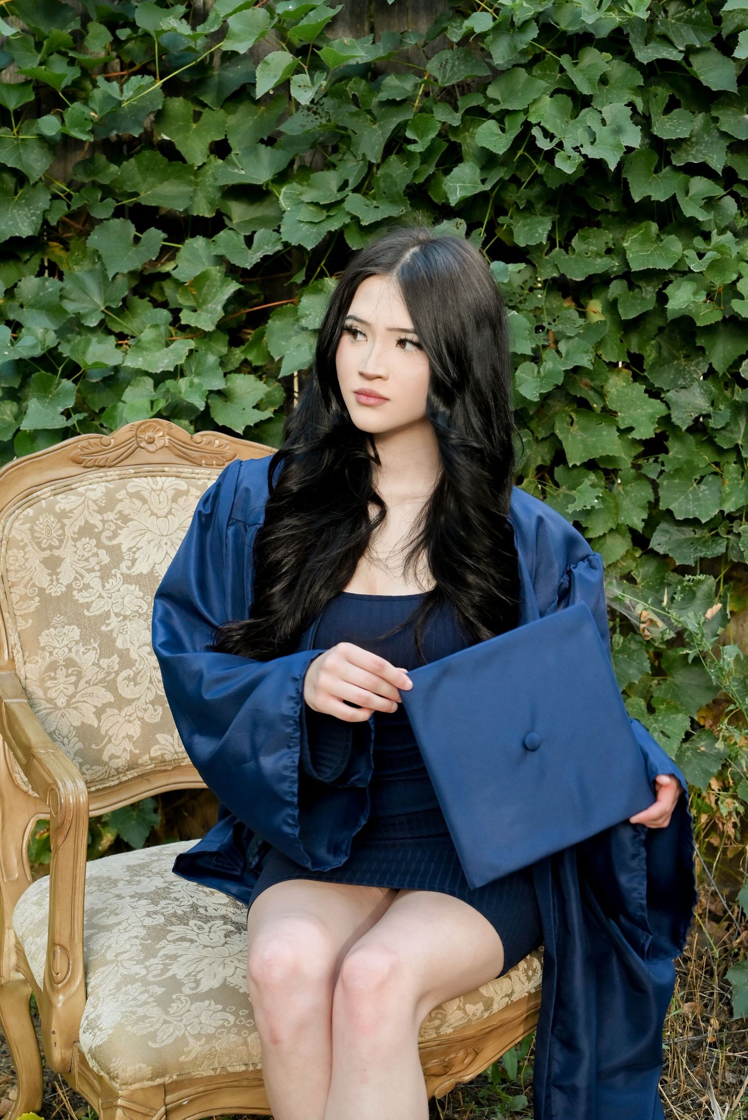 A figure in a navy blue dress sits on an ornate vintage beige sofa against a backdrop of green ivy leaves.