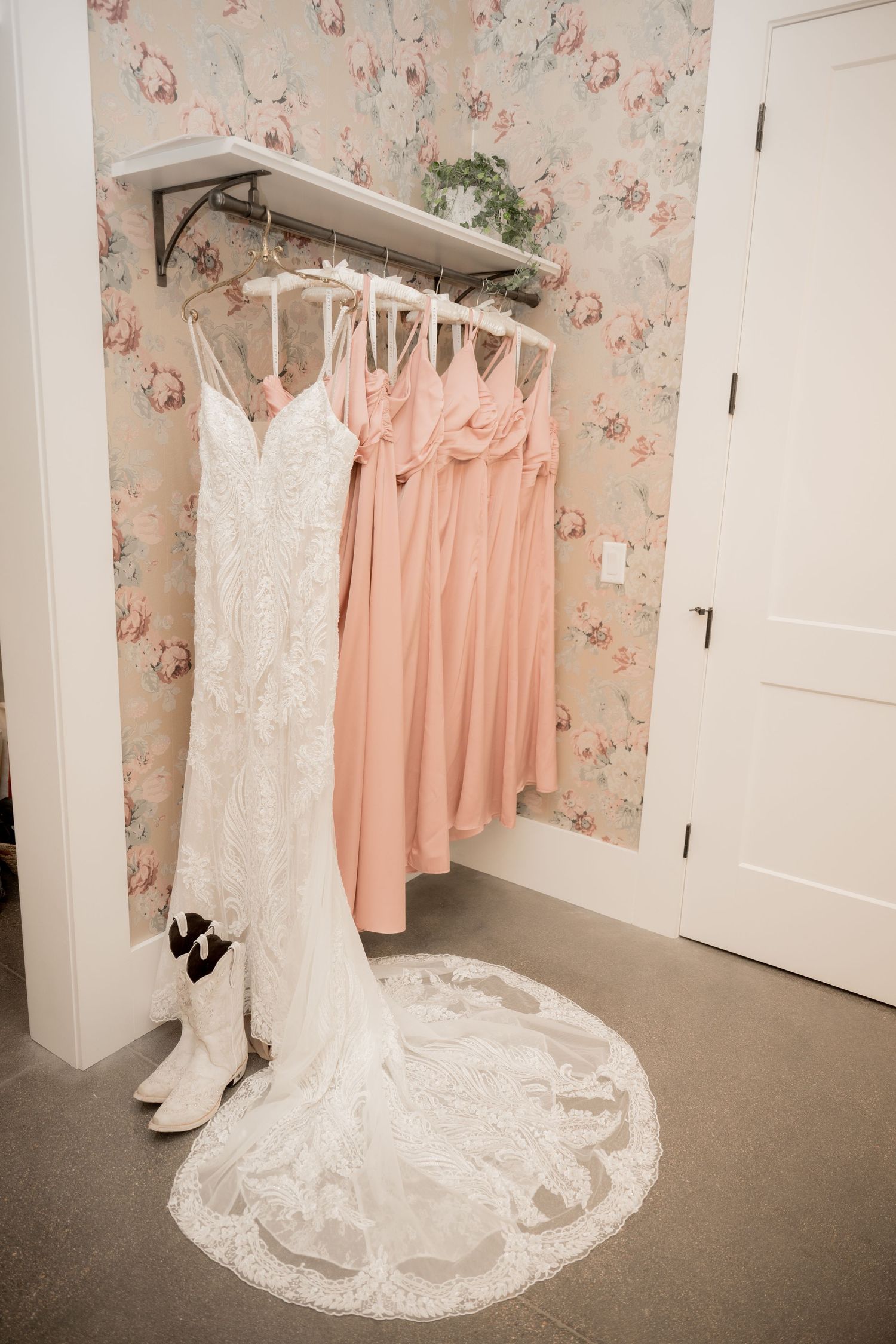 Wedding dress and pink bridesmaid dresses hang on display in a bridal boutique with floral wallpaper.