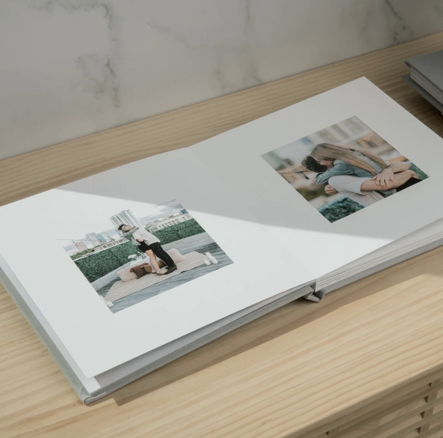 A white photo album book lies open on a wooden surface showing blank pages and photo layouts.