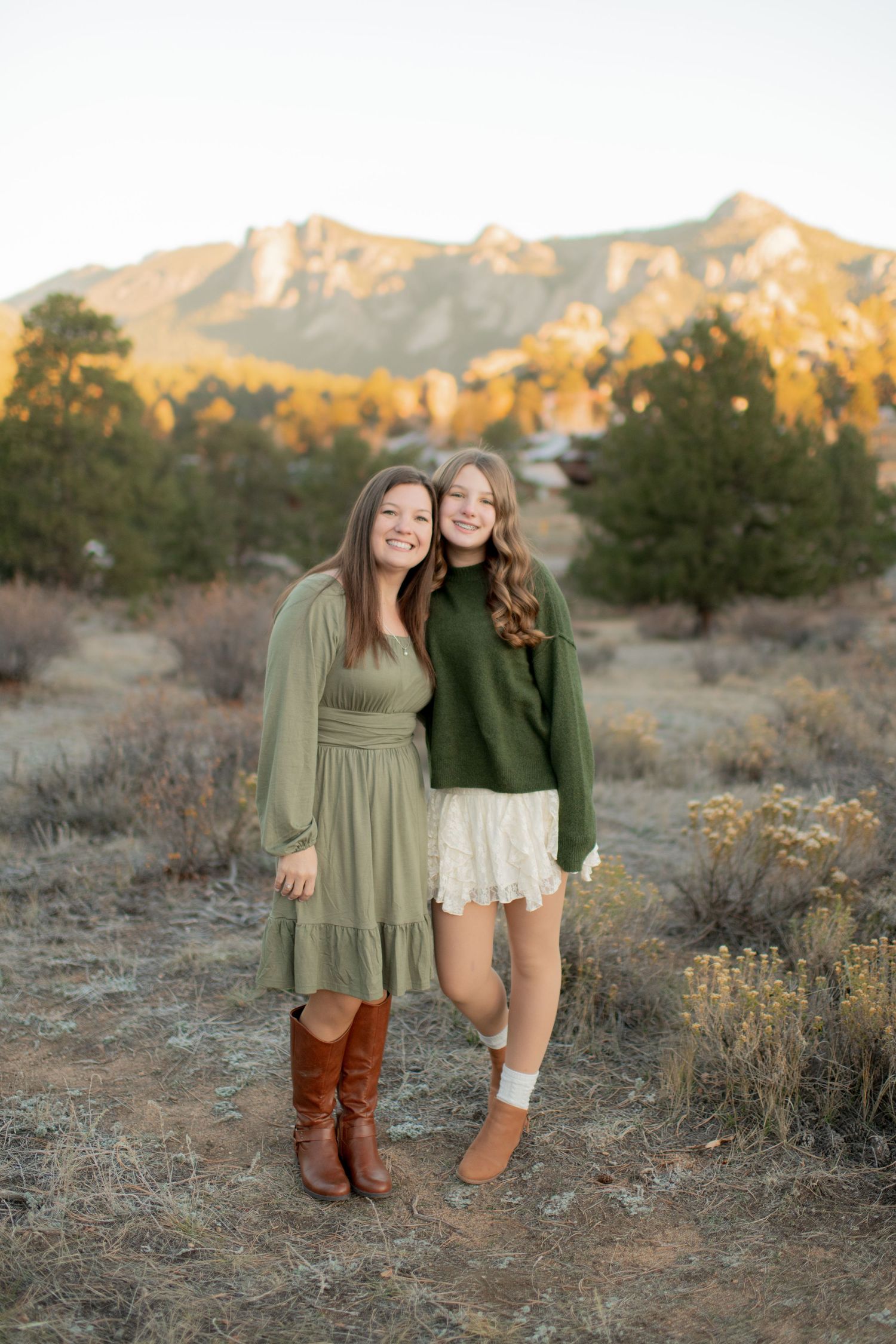 Two friends in casual dresses and boots pose together in a desert landscape with mountains at sunset.