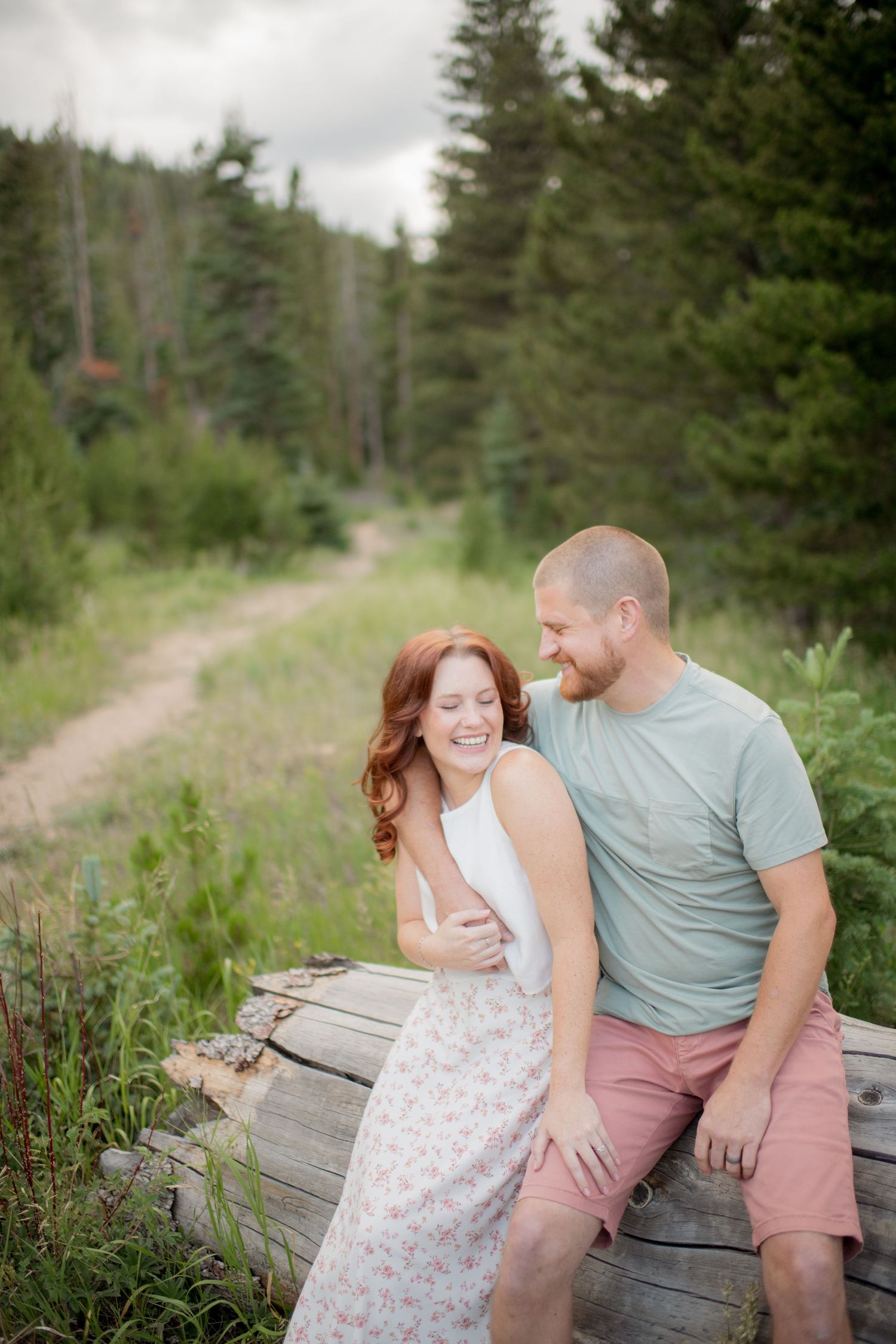 A couple shares a joyful moment while sitting on rustic logs along a forest trail on a cloudy summer day.