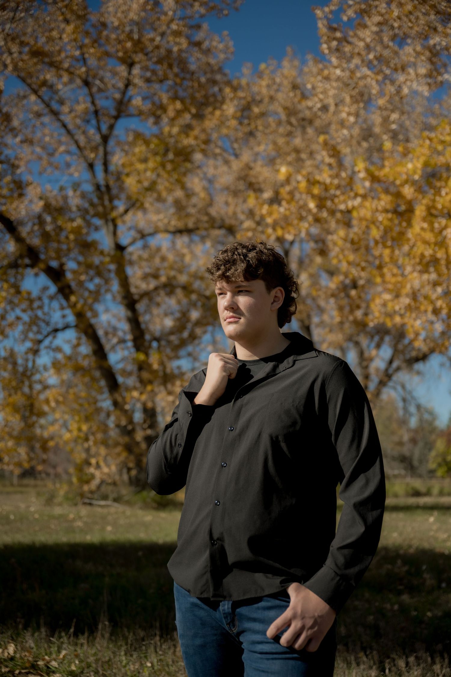 Person wearing a black hoodie and jeans stands outdoors in autumn with golden trees in the background.