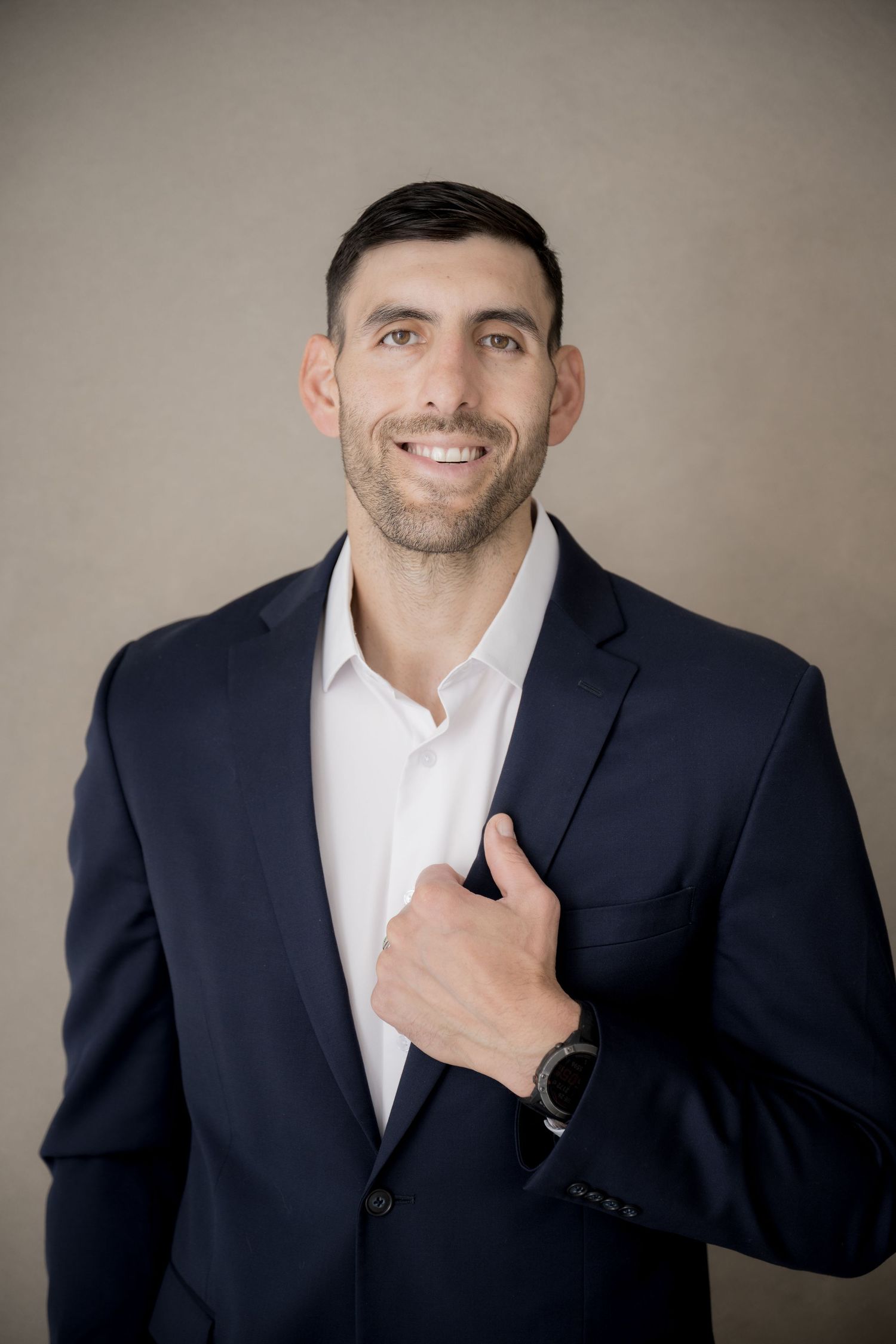 Professional headshot of a person in a navy suit and white dress shirt smiling against a neutral background.