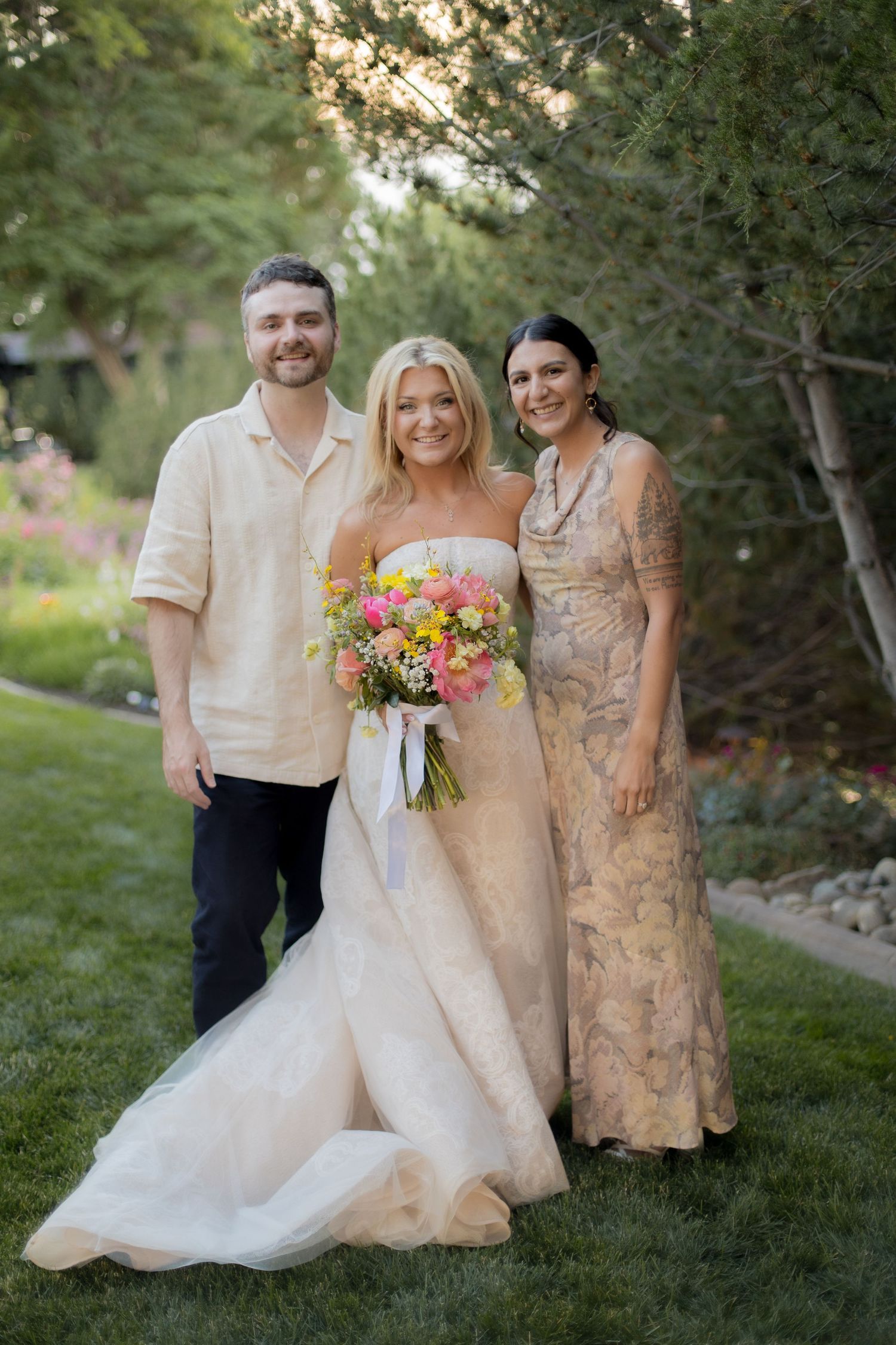 A group stands together outdoors in formal attire with colorful floral bouquets against a natural garden backdrop.