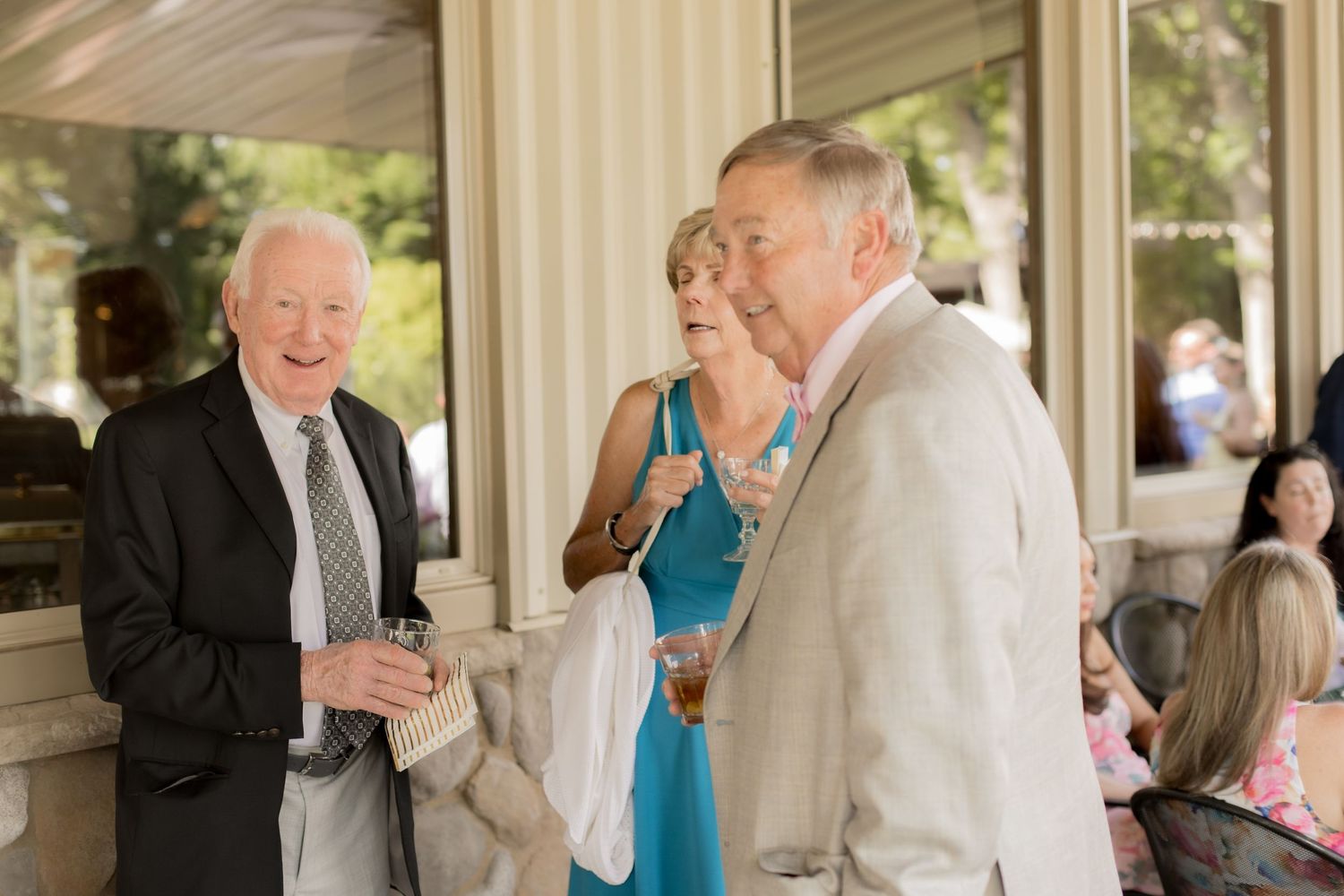 People in formal attire gather at an outdoor venue with white columns during what appears to be a social event.