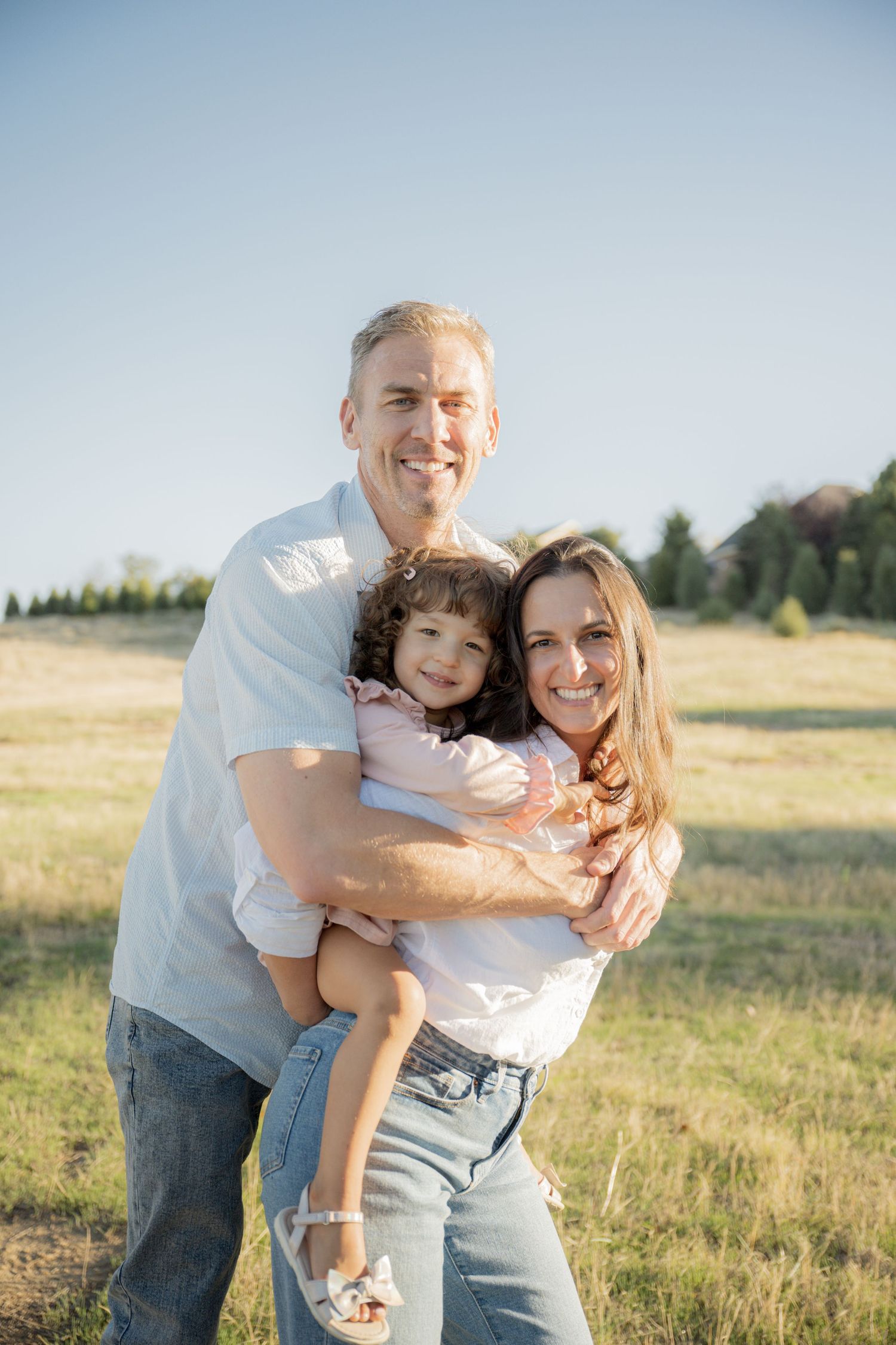 A couple embraces playfully in a sunny meadow with golden grass and trees in the background.