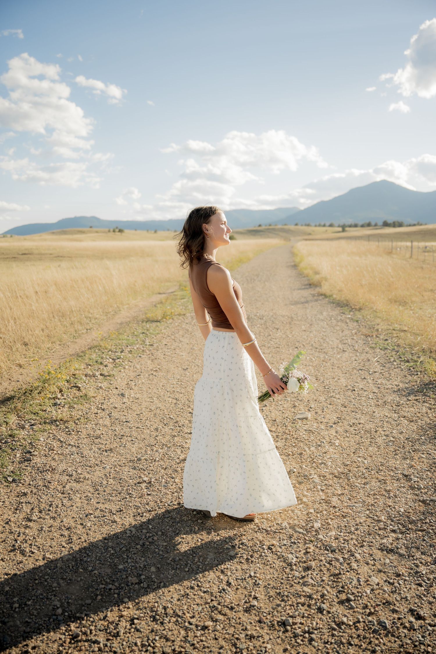 A bride in a white wedding dress stands on a gravel road with mountains and golden fields in the distance.