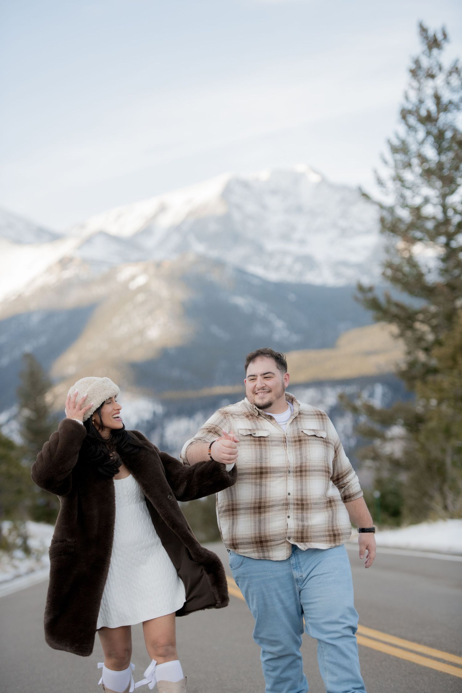 Two people dance playfully on a mountain road with snow-capped peaks visible in the background.