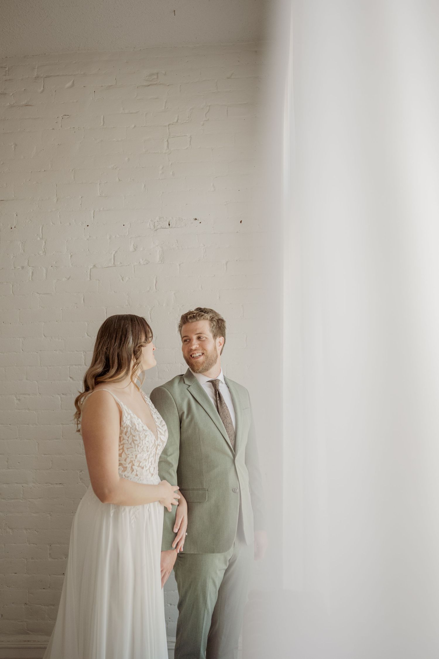 A couple in wedding attire share an intimate moment against a white wall in a minimalist setting.