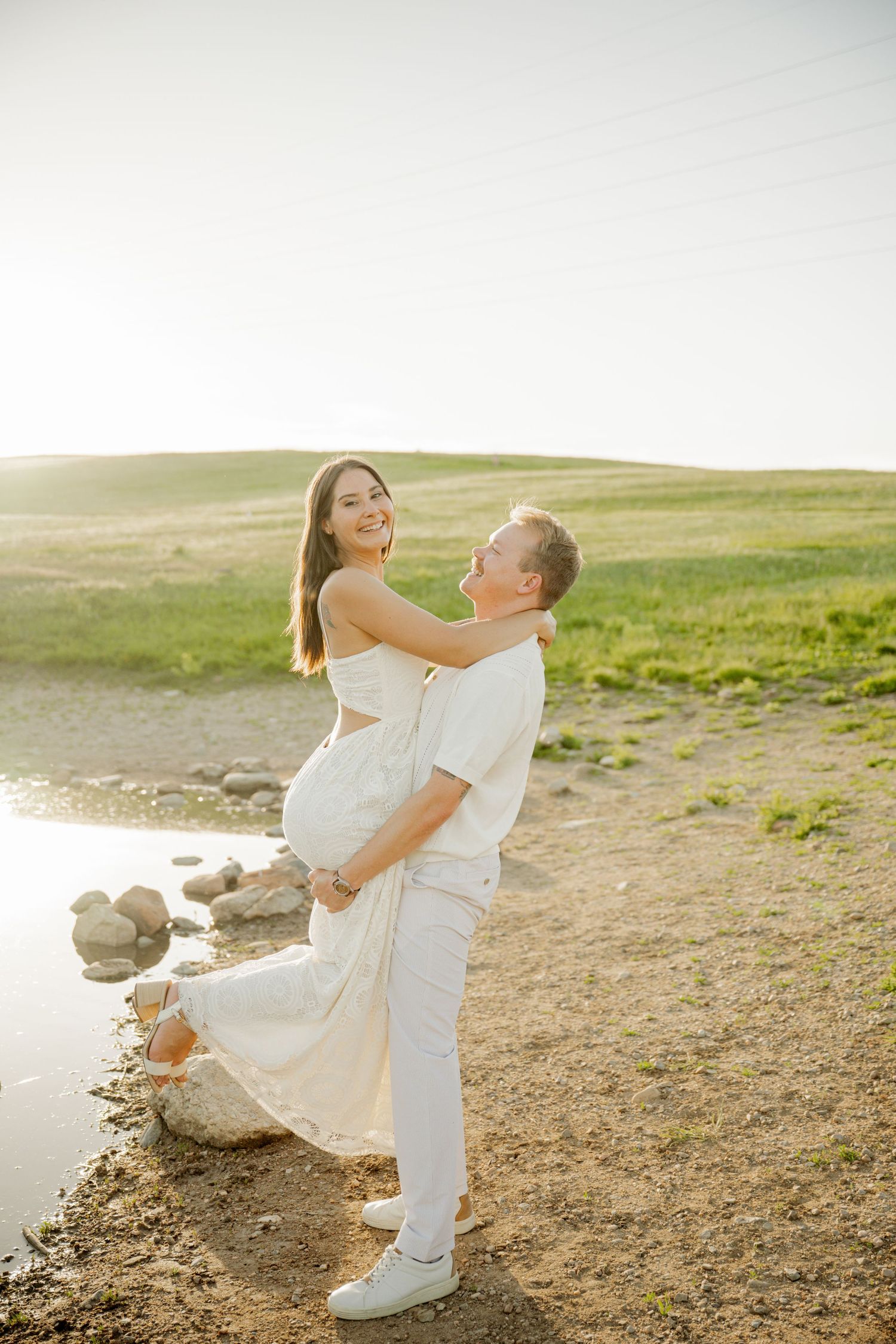 A loving couple in white outfits share a romantic moment along a shoreline with rolling hills in the background.