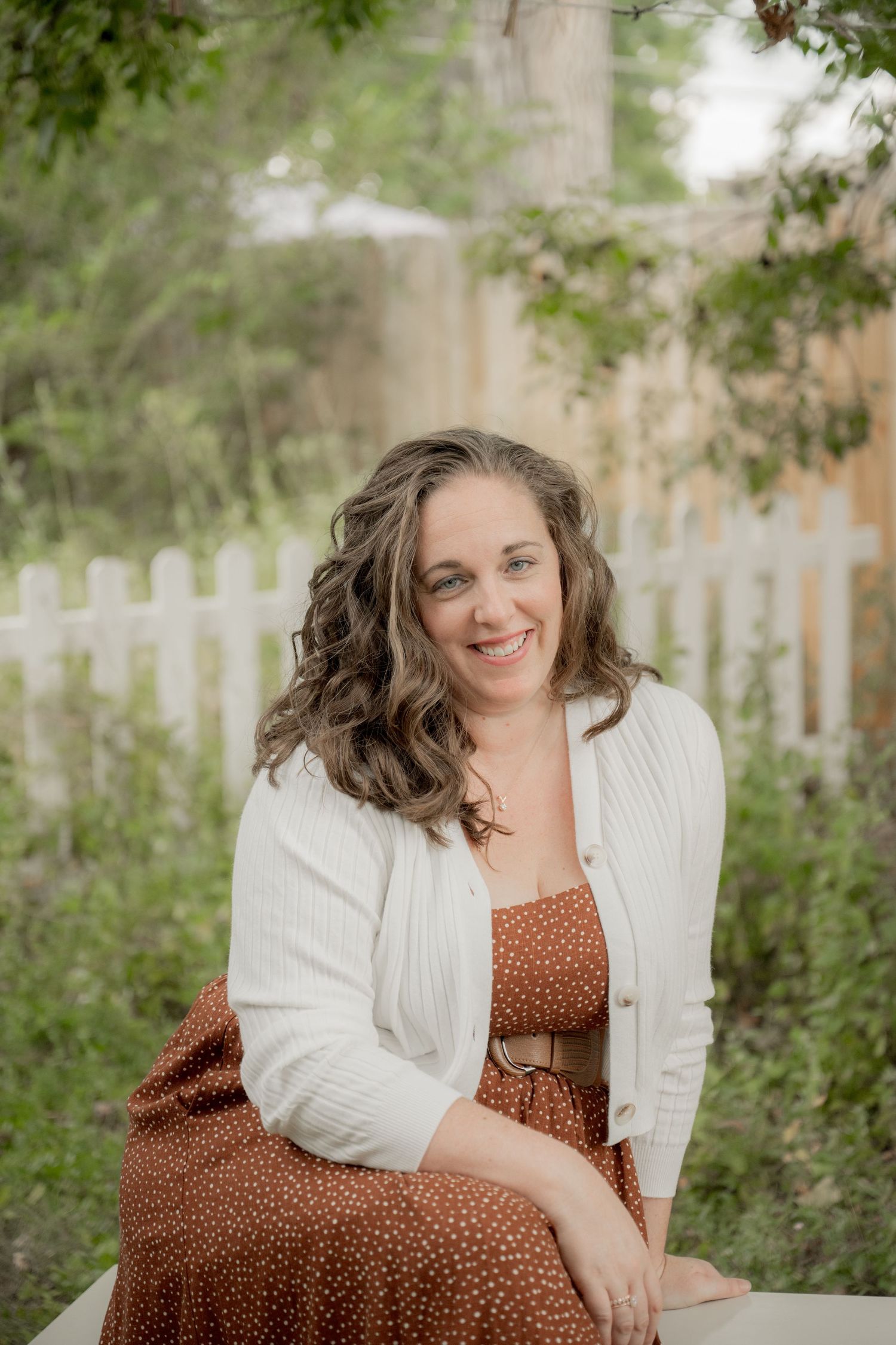 Outdoor portrait in garden setting with white picket fence, featuring person in brown polka dot dress and white cardigan.