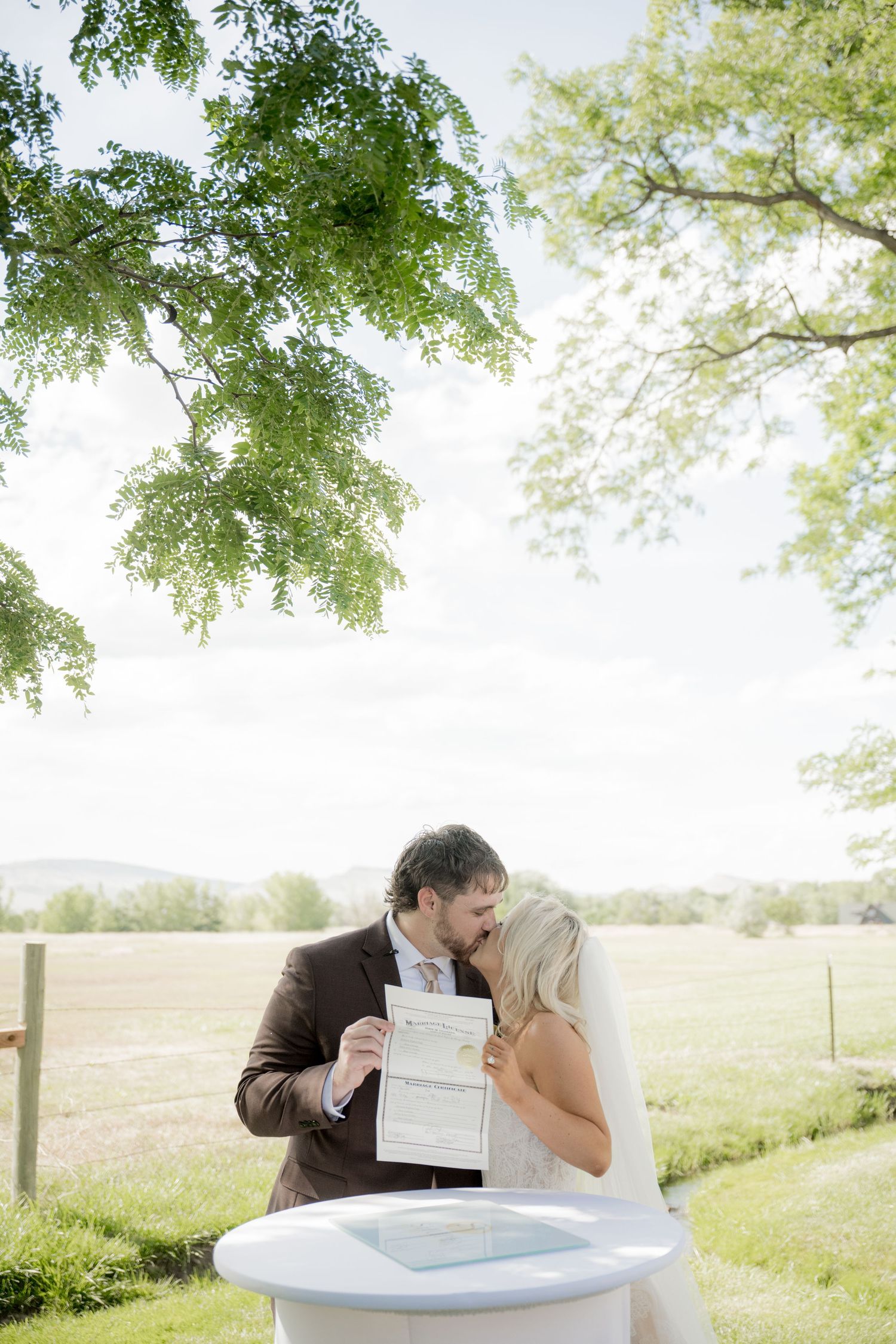 A couple shares a kiss while holding their marriage certificate outdoors on a bright spring day in a pastoral setting.