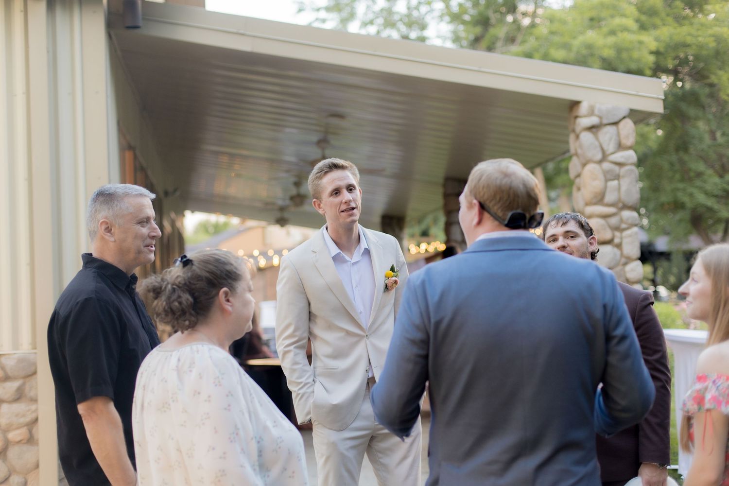 Wedding guests gather at an outdoor reception venue with stone pillars and greenery in the background.