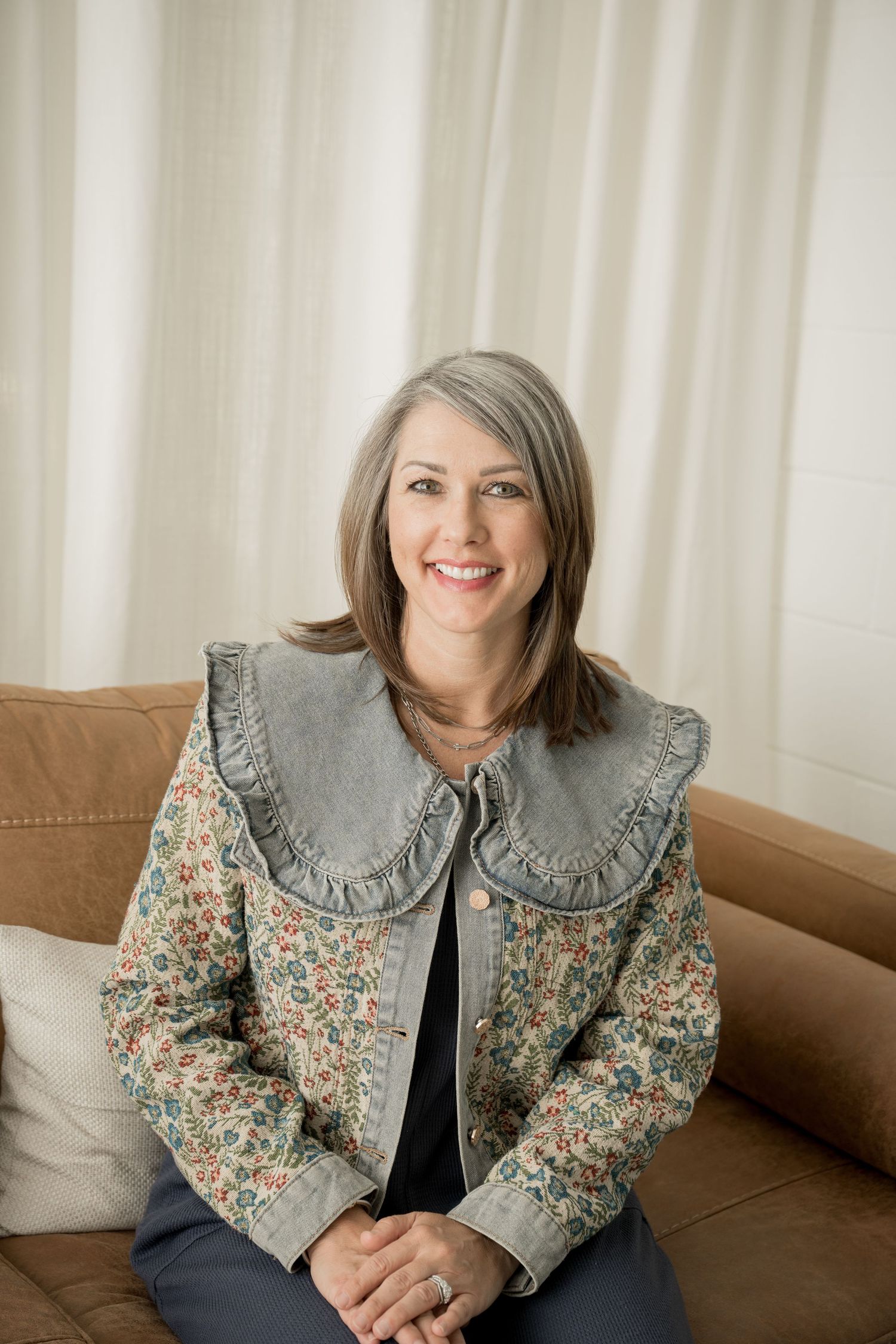 A woman wearing a floral print jacket with a gray ruffled collar sits on a brown couch in a bright room.