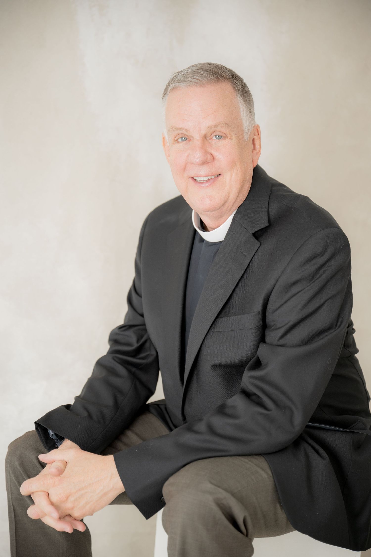 Professional studio portrait of a person wearing a black suit jacket and gray sweater seated against a light background.