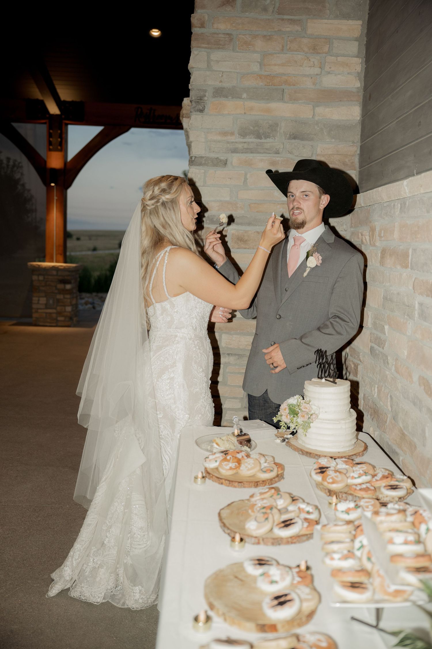 A bride in a white lace gown feeds wedding cake to her groom in a grey suit and cowboy hat at their rustic reception.