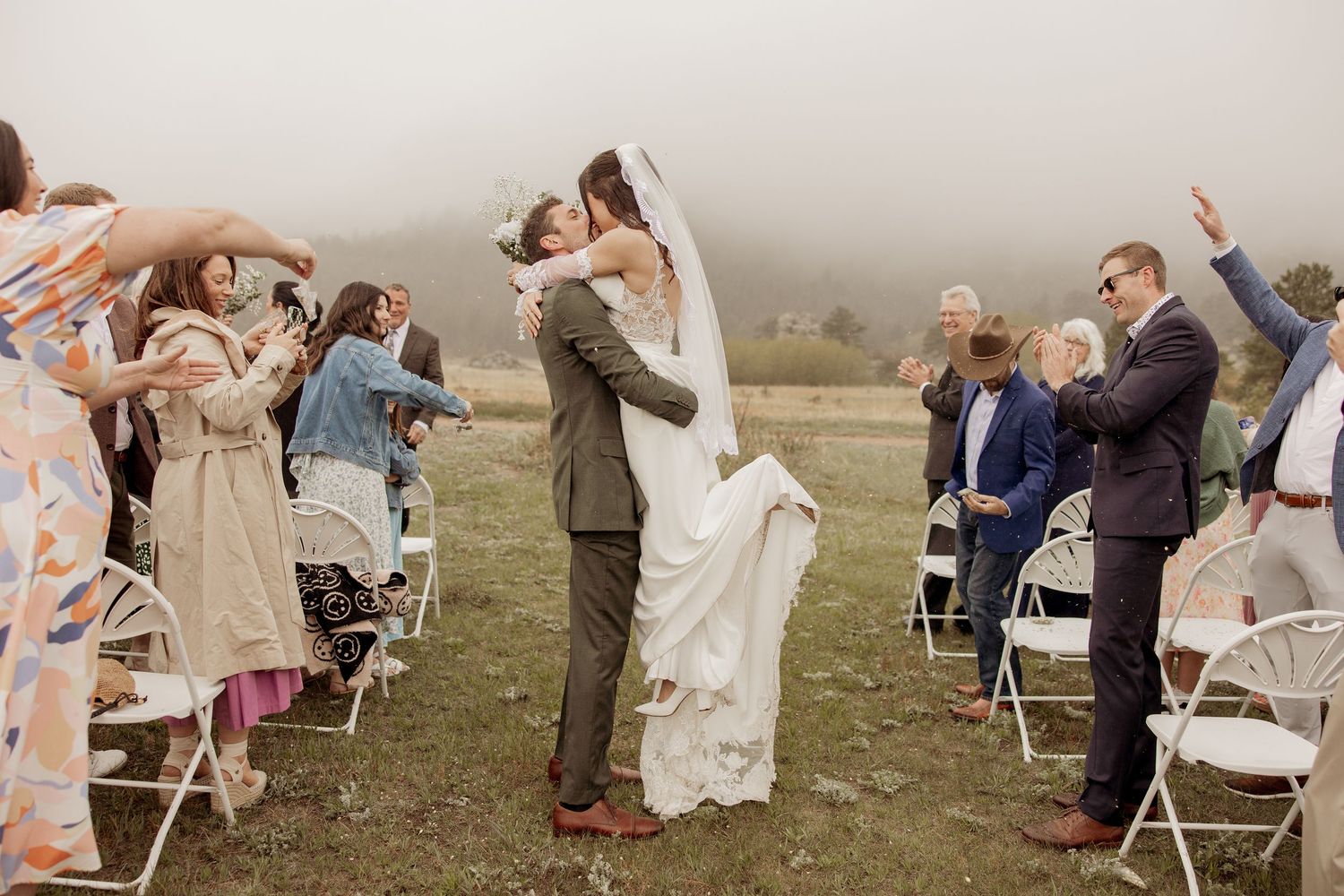 A wedding couple shares their first kiss at a misty outdoor ceremony as guests celebrate with raised hands.