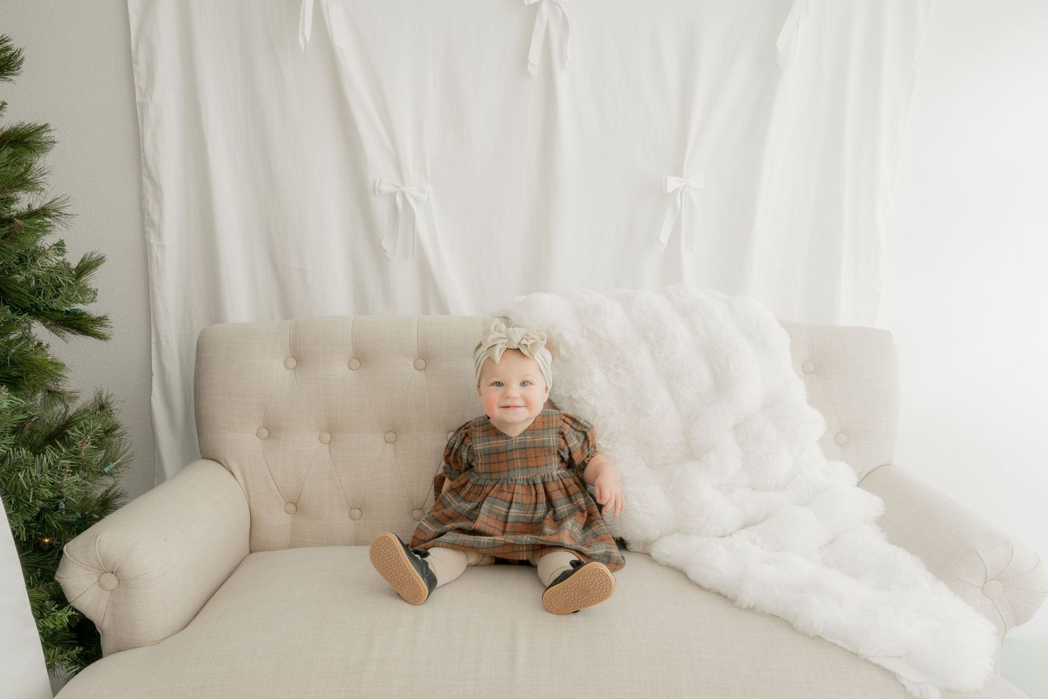 A white tufted bench with decorative pillows sits against a white curtain backdrop near green foliage.