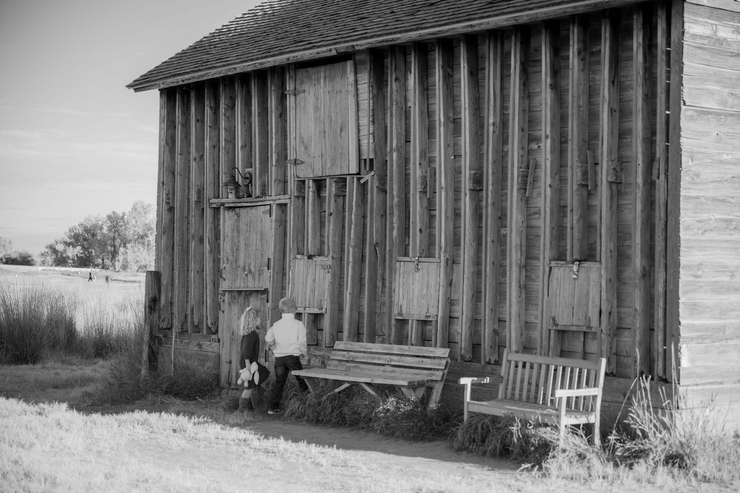 A rustic wooden barn with weathered vertical boards and two wooden benches sitting outside in black and white.
