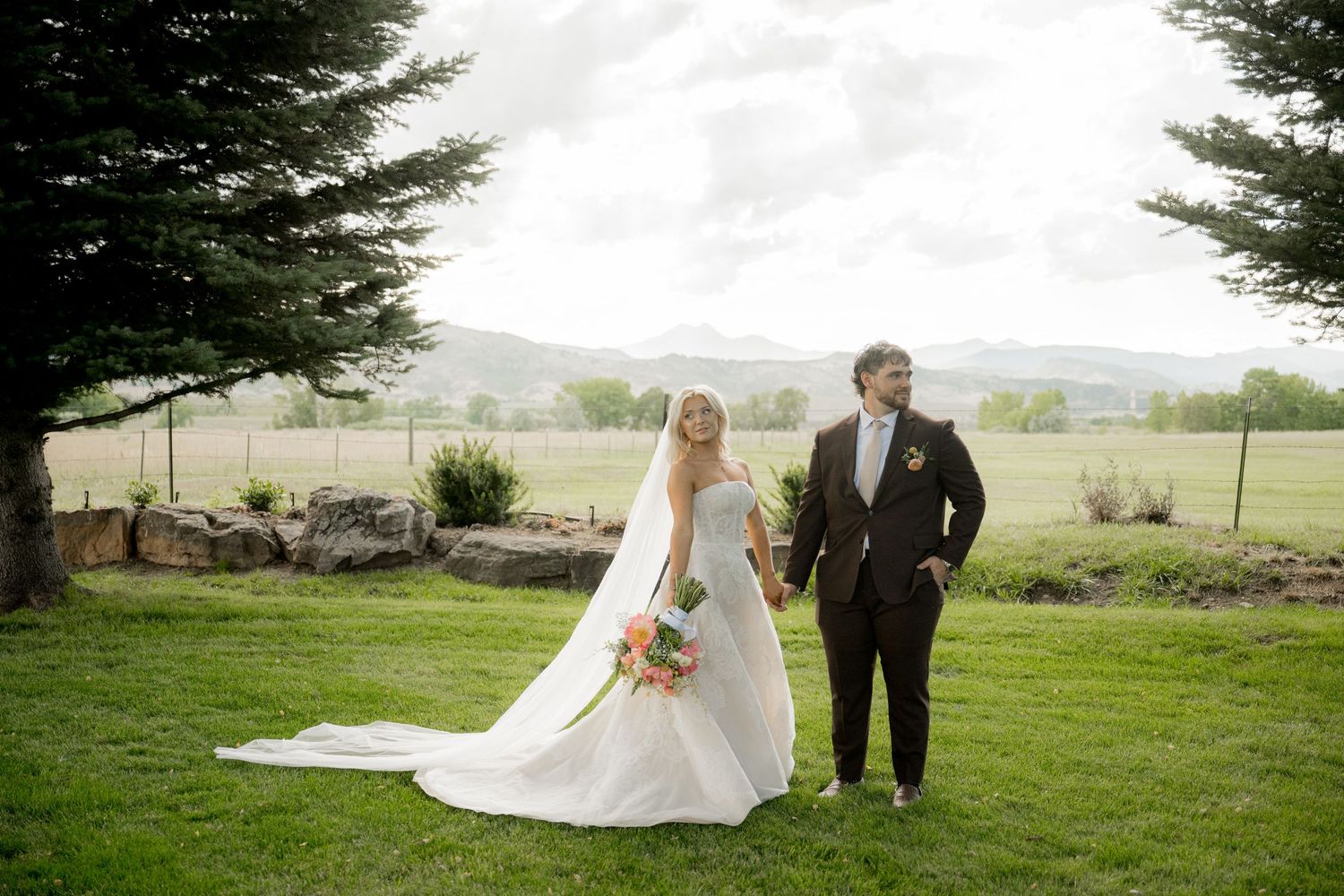 Wedding couple poses in front of evergreen trees on a scenic grassy hillside with mountains in the distance.