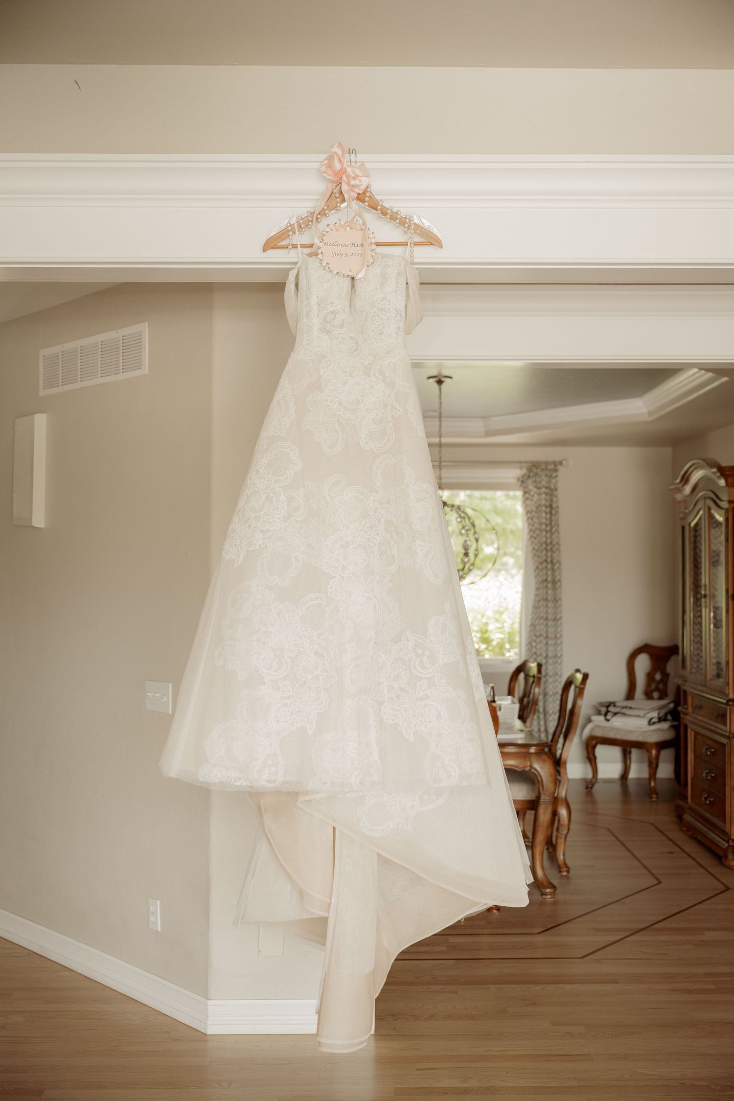 A wedding dress hangs elegantly in a sunlit room with hardwood floors and white walls.