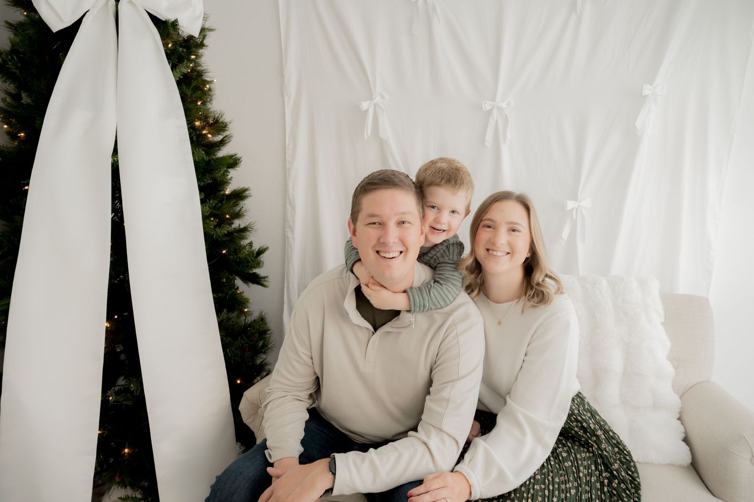 Family wearing neutral colored sweaters poses together in front of white backdrop with Christmas tree.