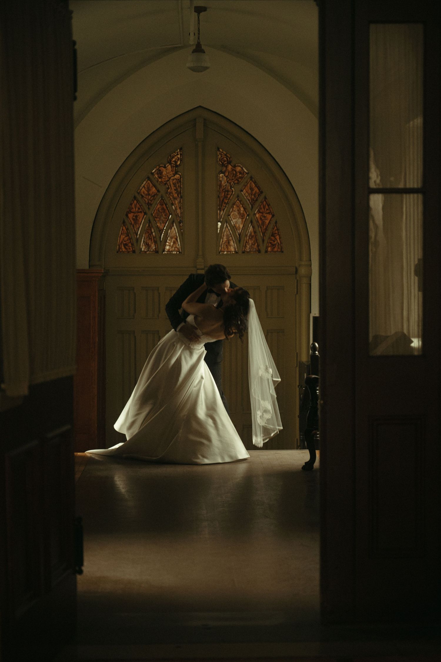 Bride and groom sharing a romantic kiss in a dimly lit church with stunning stained glass windows.