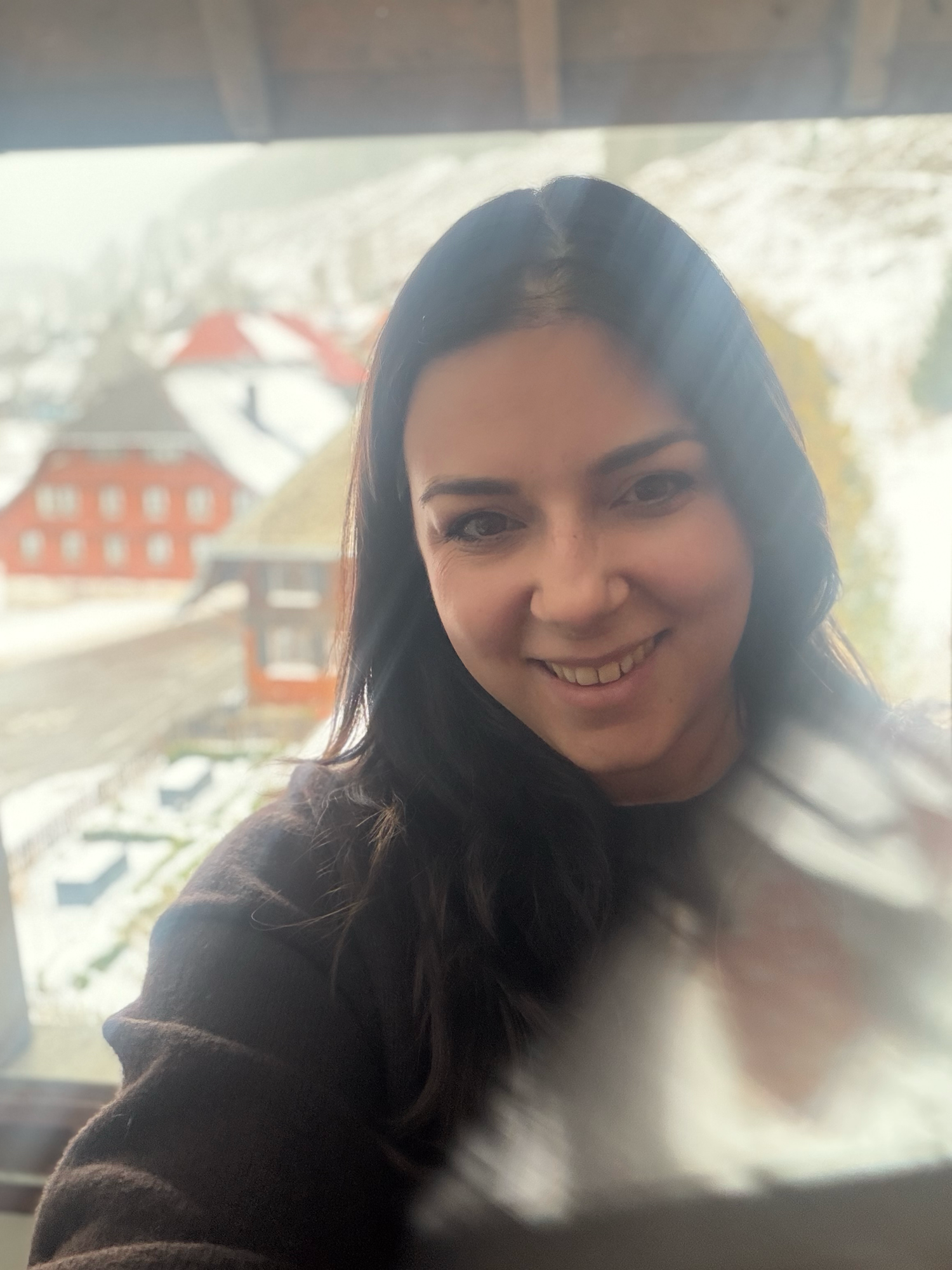 A person with long dark hair smiles warmly while sitting near a window with buildings visible outside.