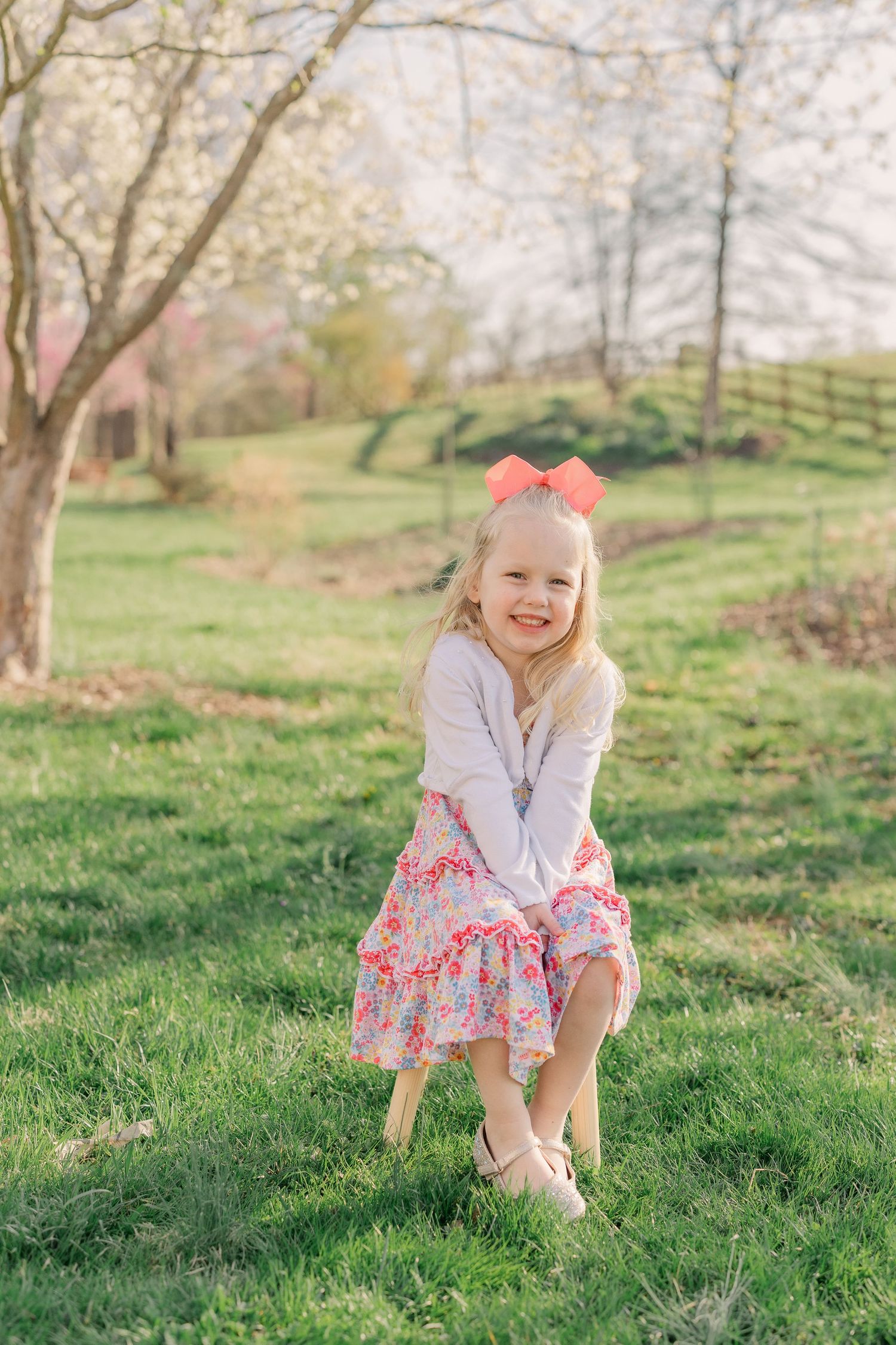 A child in a pink floral dress and bow plays on green grass during springtime with blooming trees in the background.